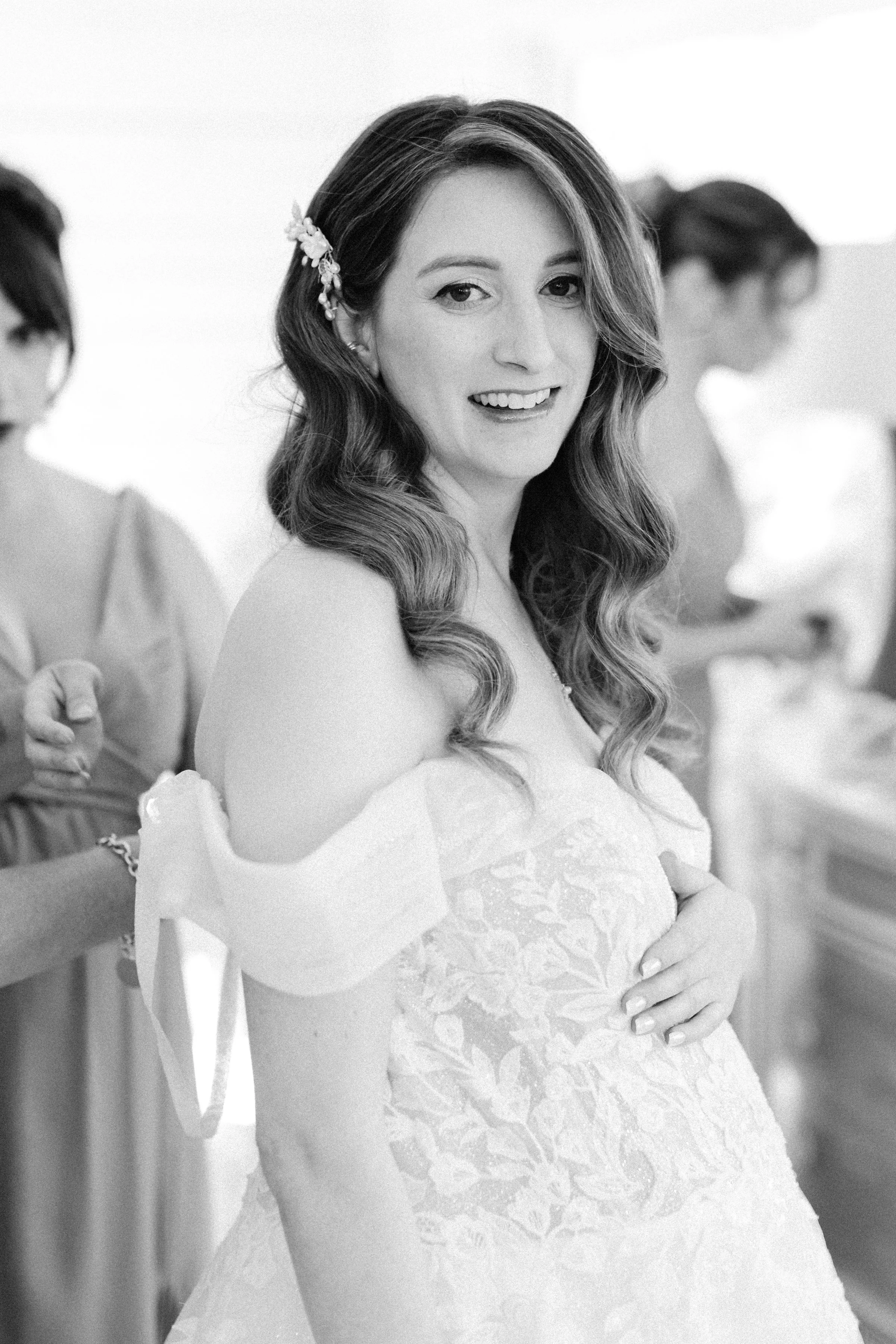 Black and white photo of a smiling woman in a wedding dress, with long wavy hair and an ornate hair accessory, holding her hand on her belly, surrounded by other women preparing for a wedding.