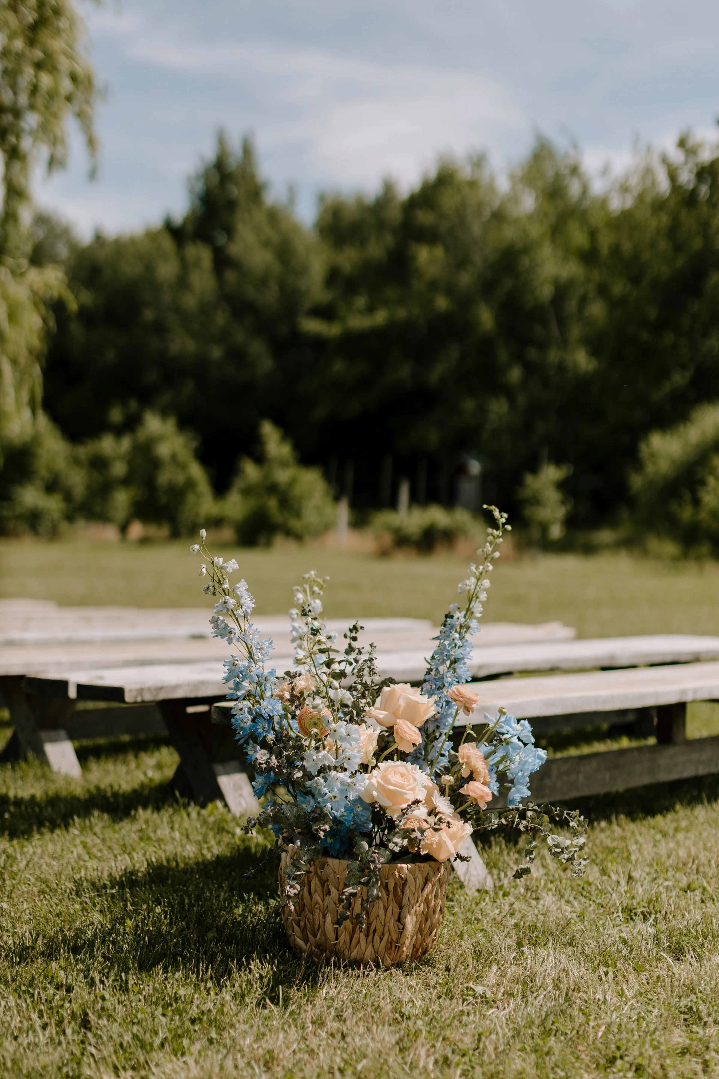 A bouquet of pastel-colored flowers in a woven basket, placed on the grass near a picnic table outdoors with trees and a cloudy sky in the background.