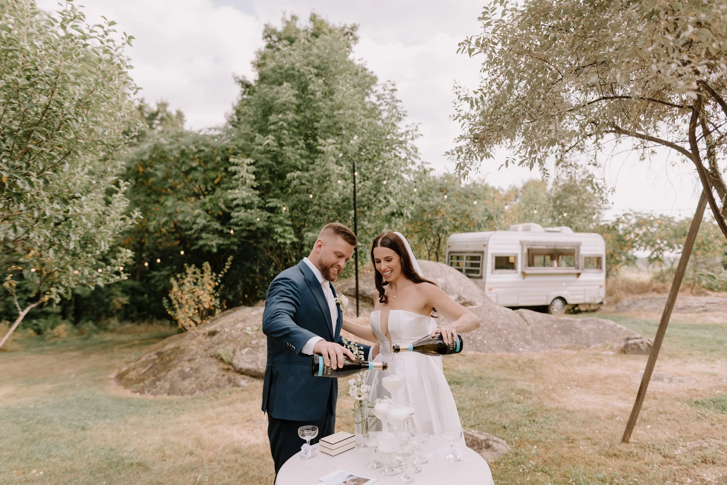 A newlywed couple, a man in a suit and a woman in a white wedding dress, are celebrating outdoors with a champagne tower on a table. The man is holding a champagne bottle and pouring into glasses, smiling. The background features green trees, a cloud