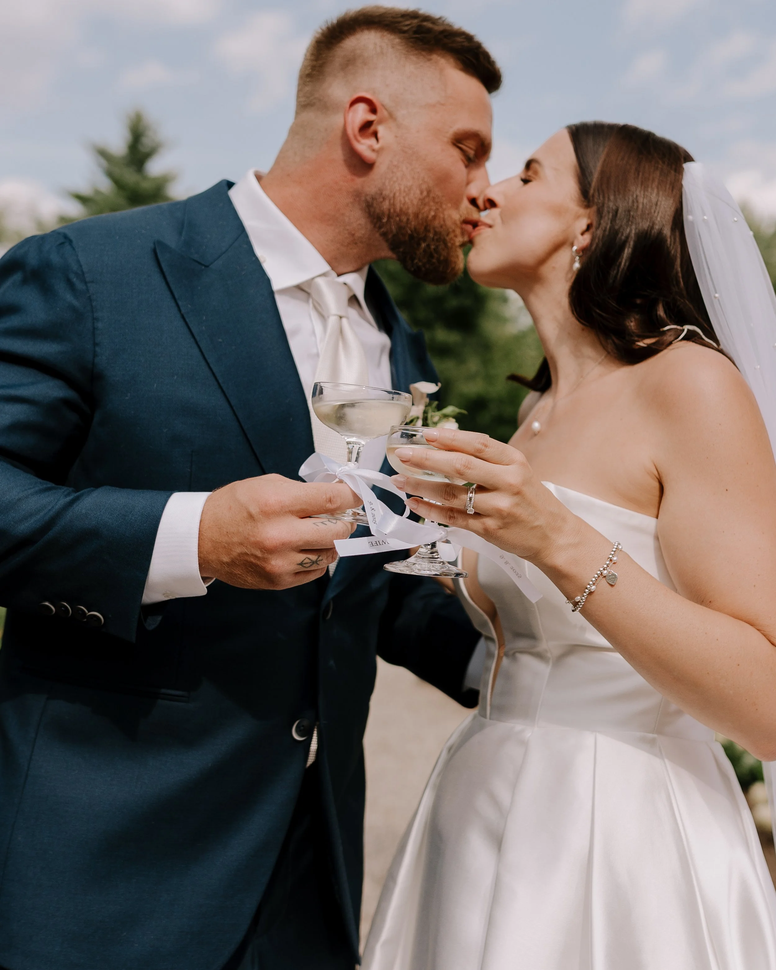 A bride and groom sharing a kiss during their outdoor wedding ceremony, holding champagne glasses.