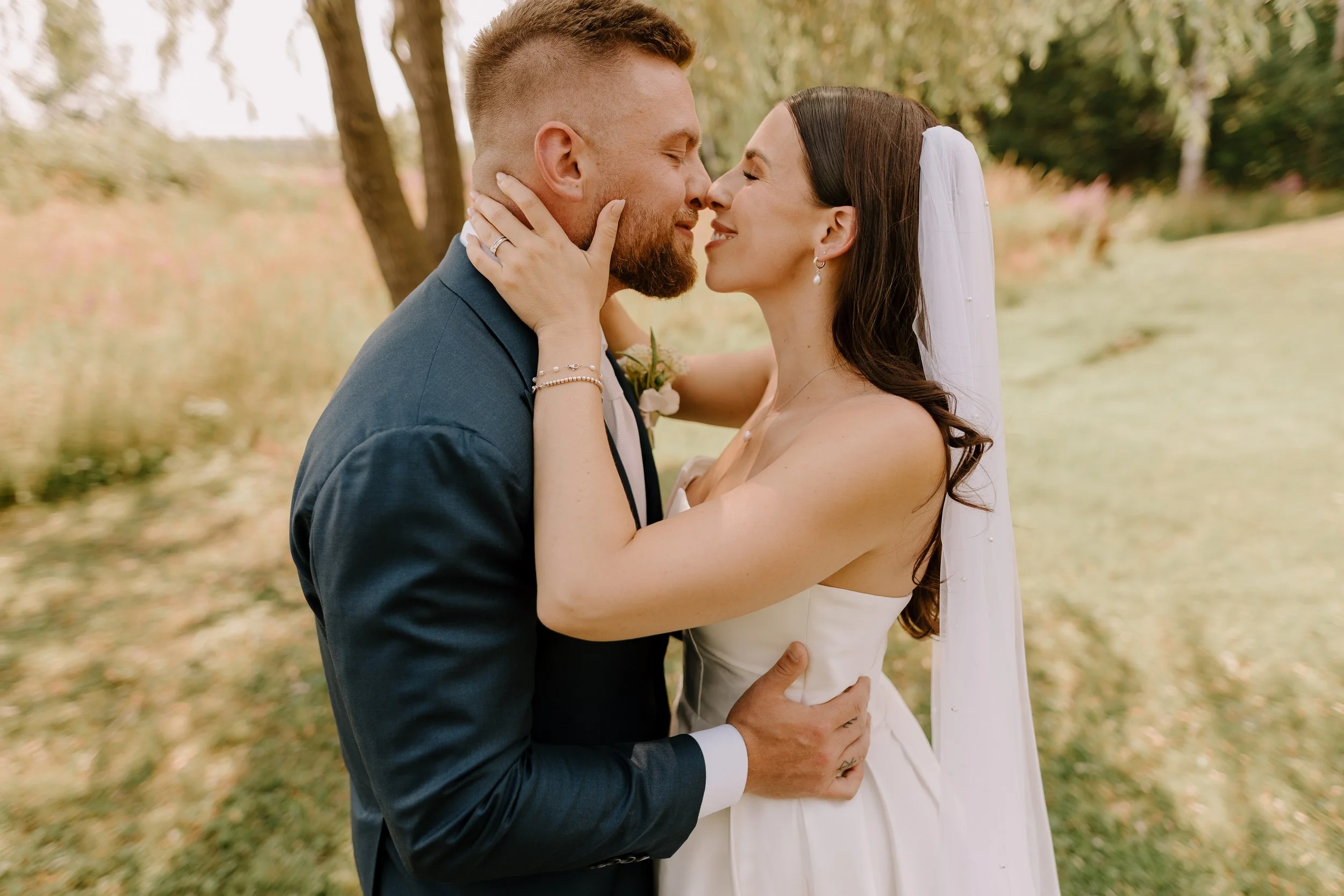 A bride and groom share a romantic moment outdoors, embracing and about to kiss, with trees and fallen leaves in the background.