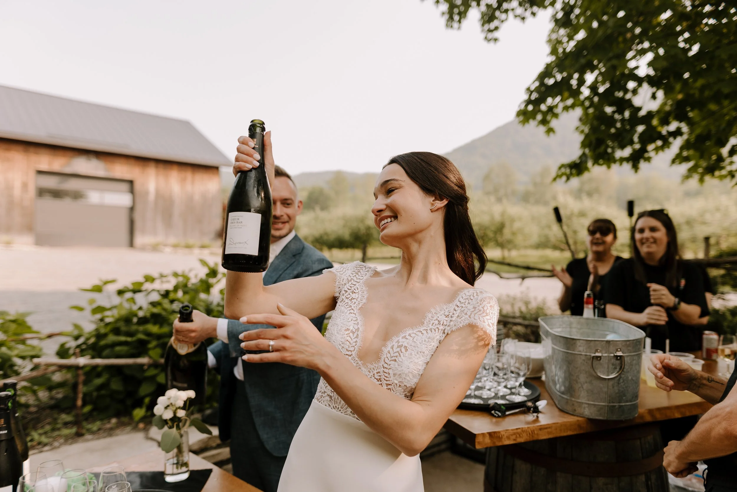 A woman in a white lace dress holding a wine bottle at an outdoor gathering, smiling as she joins others behind a table with glasses and a bucket of ice, with a barn and trees in the background.