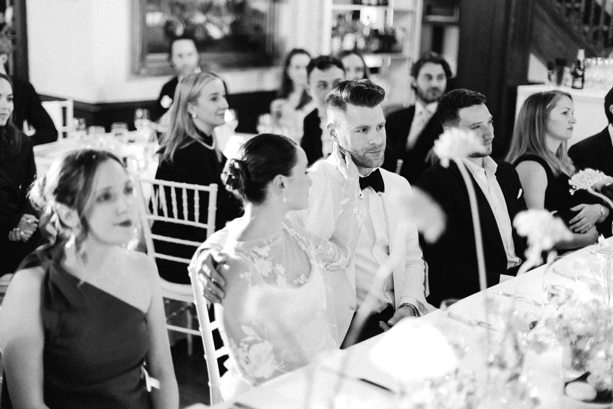 A black-and-white photo of a formal gathering with people seated at tables, paying attention to an event or speaker. Some individuals are dressed in formal attire, including tuxedos and elegant dresses.