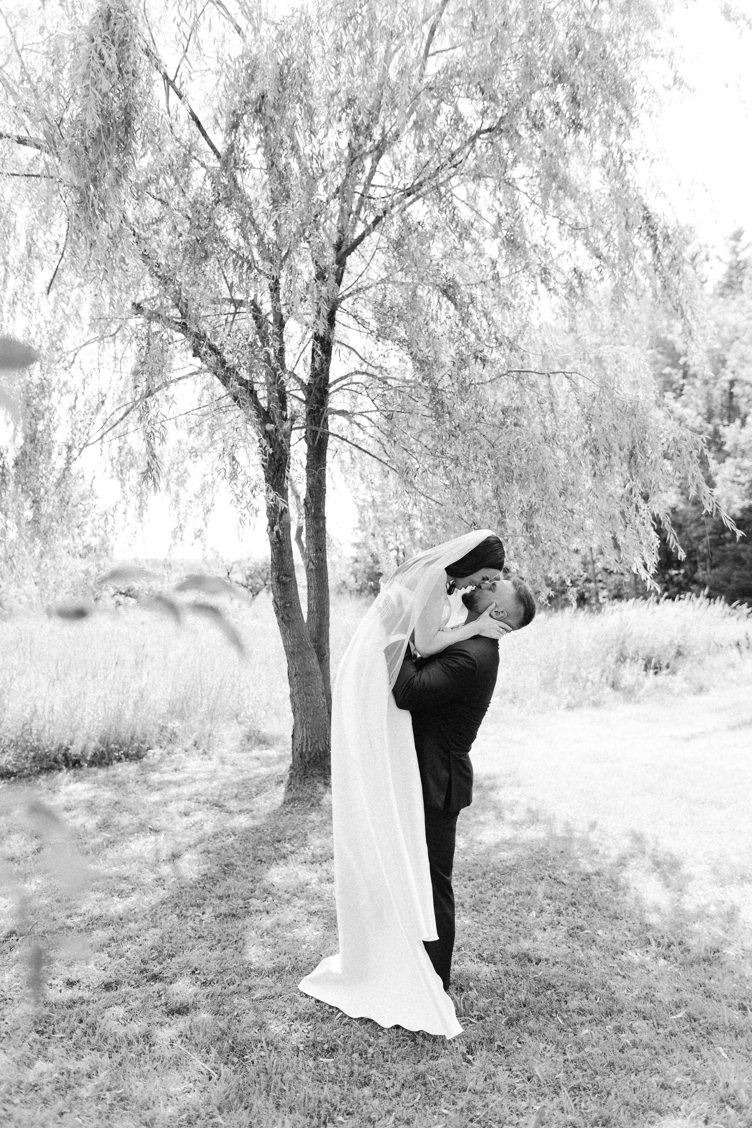 A black and white photo of a man in a suit lifting a woman in a wedding dress under a large tree in an outdoor setting. The woman is leaning toward the man, and they are sharing an intimate moment.