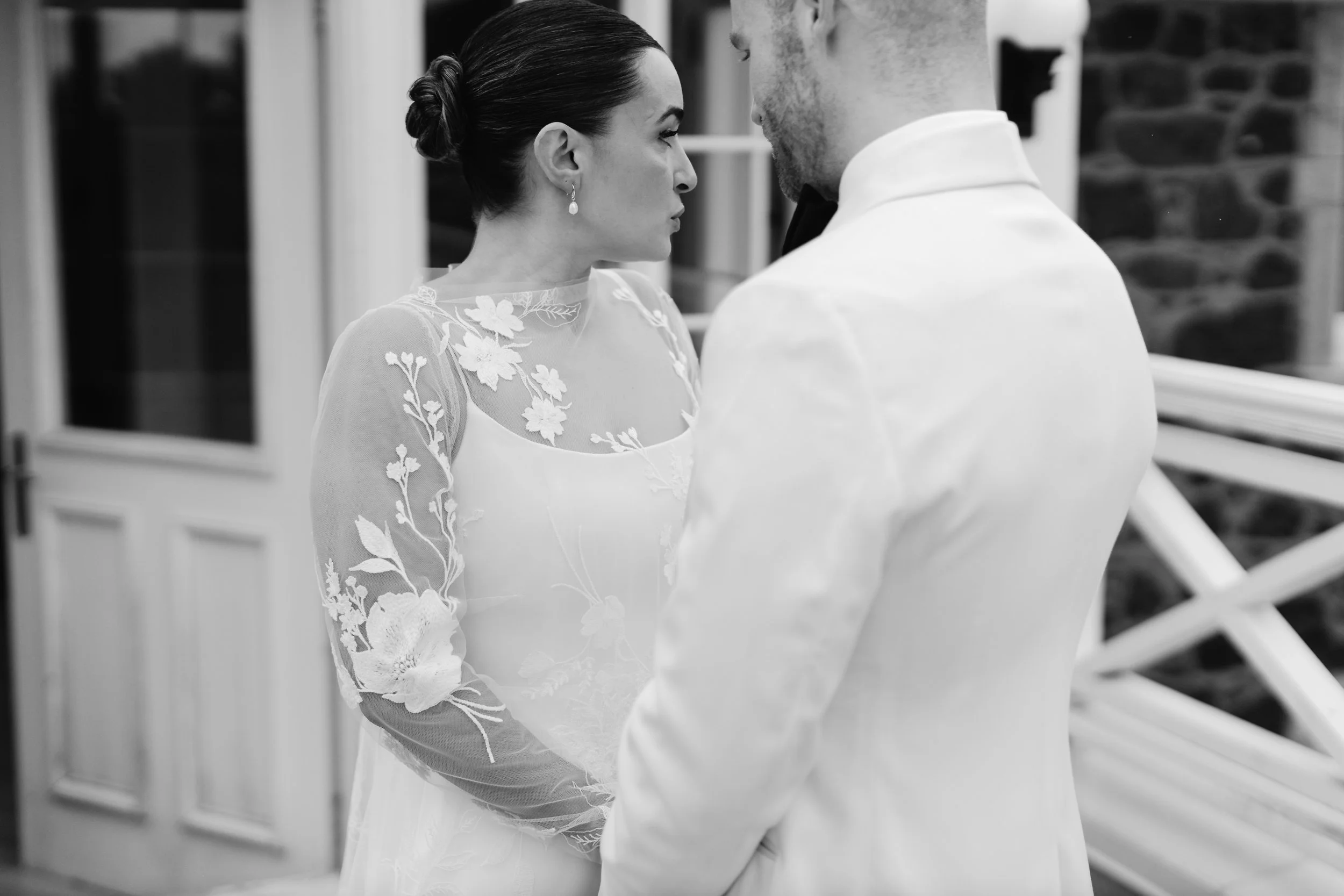 A black and white photo of a bride and groom standing close with their foreheads touching, indoors near a window and brick wall, with the bride wearing a dress with floral embroidery and pearl earrings, and the groom in a white suit.