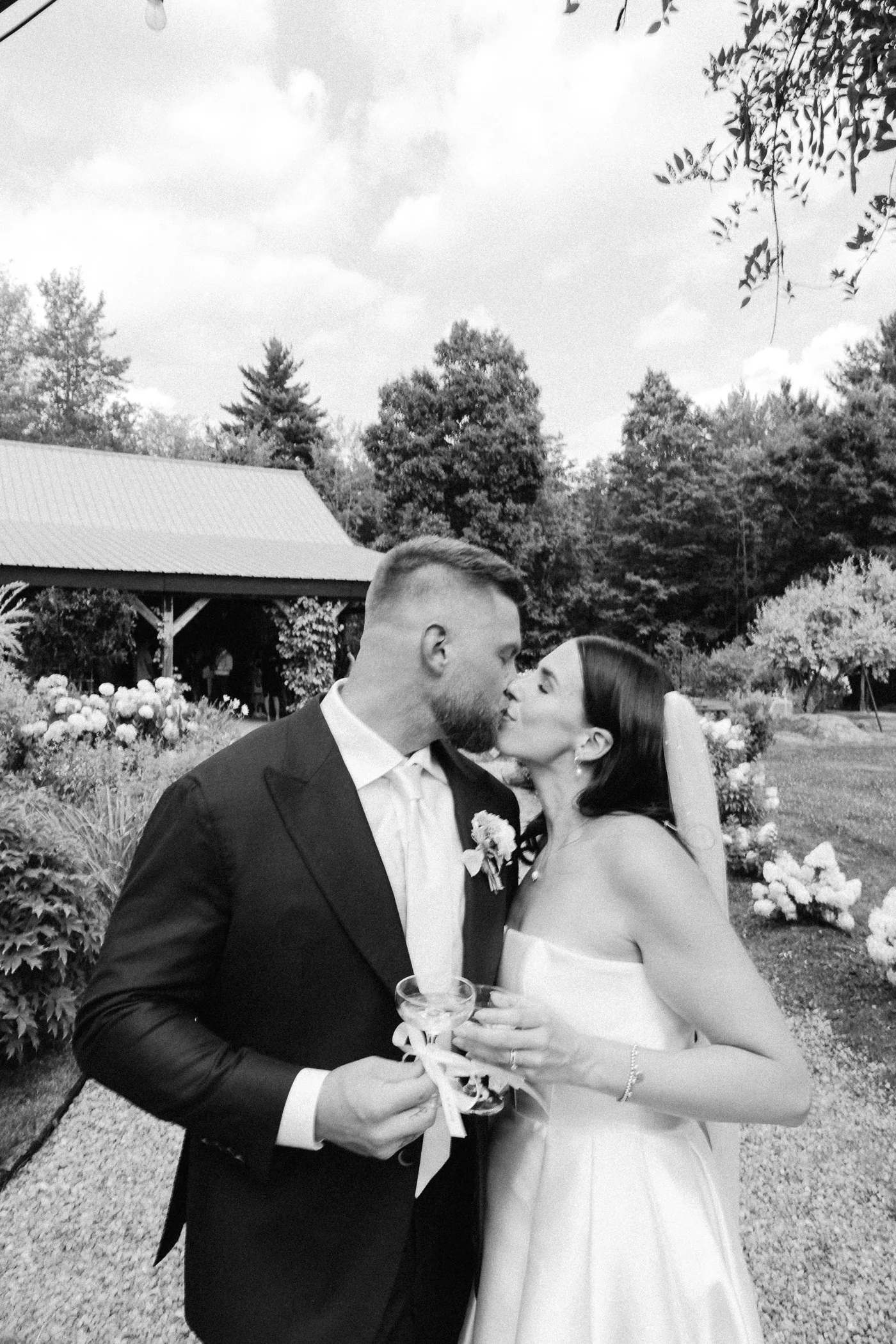 A man and woman, dressed in wedding attire, holding glasses and kissing outdoors in a garden setting with trees, flowers, and a building with a metal roof in the background.