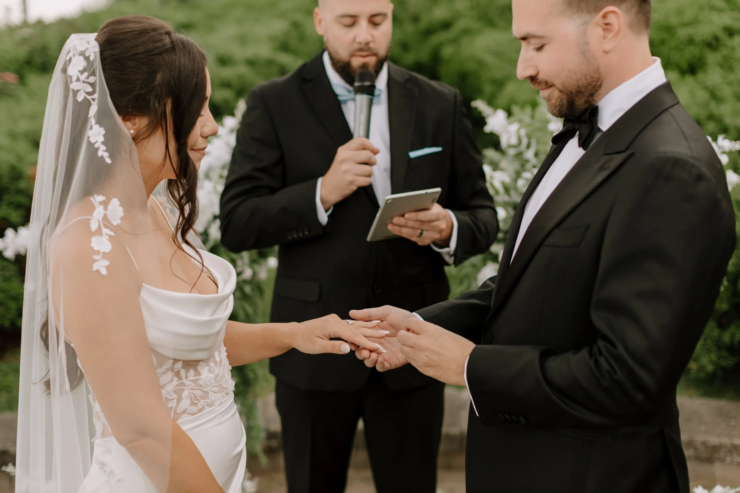 A bride and groom holding hands during their wedding ceremony, with an officiant reading vows from a tablet in the background in an outdoor setting.