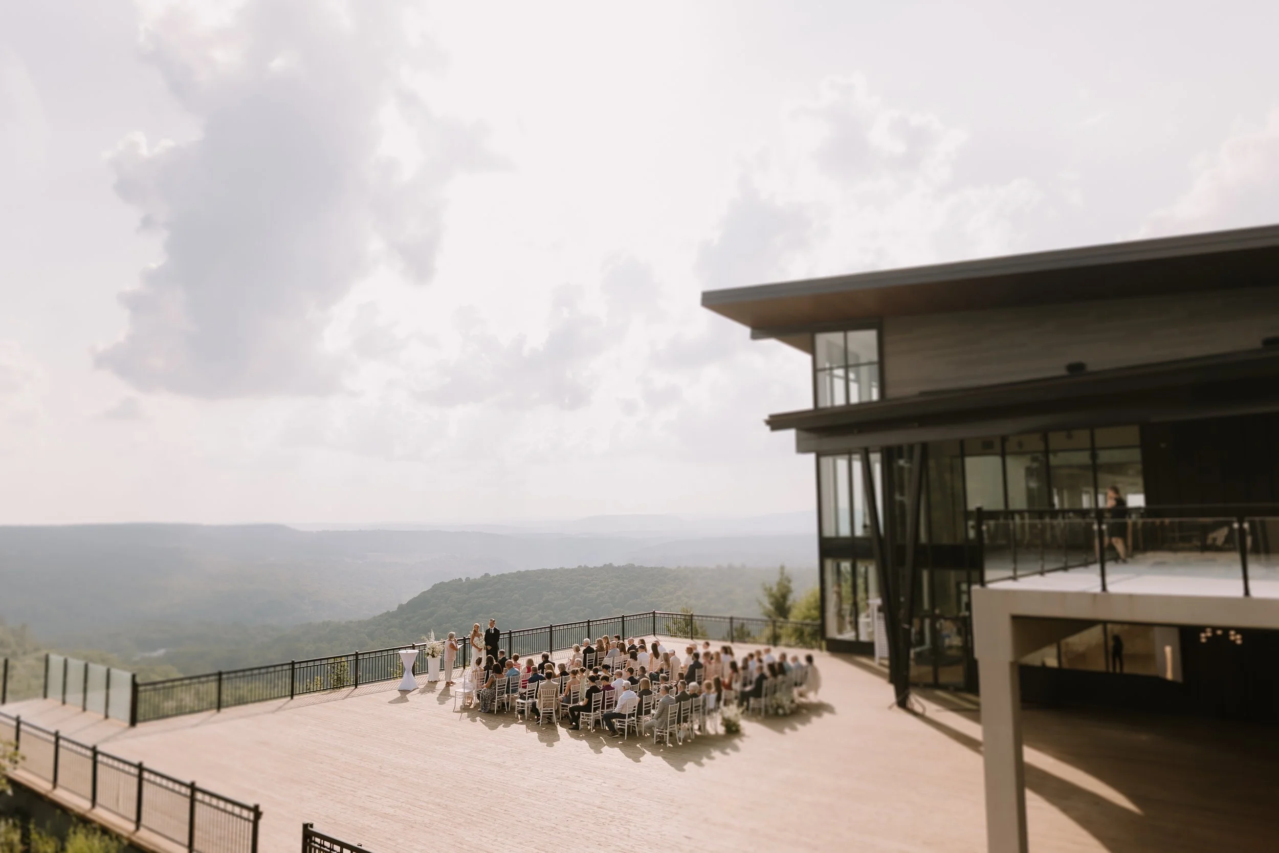 An outdoor wedding ceremony on a spacious patio with scenic mountain and lake views. People are seated in rows of white chairs, and the officiant stands before the couple exchanging vows. The modern building features large glass windows and a flat ro