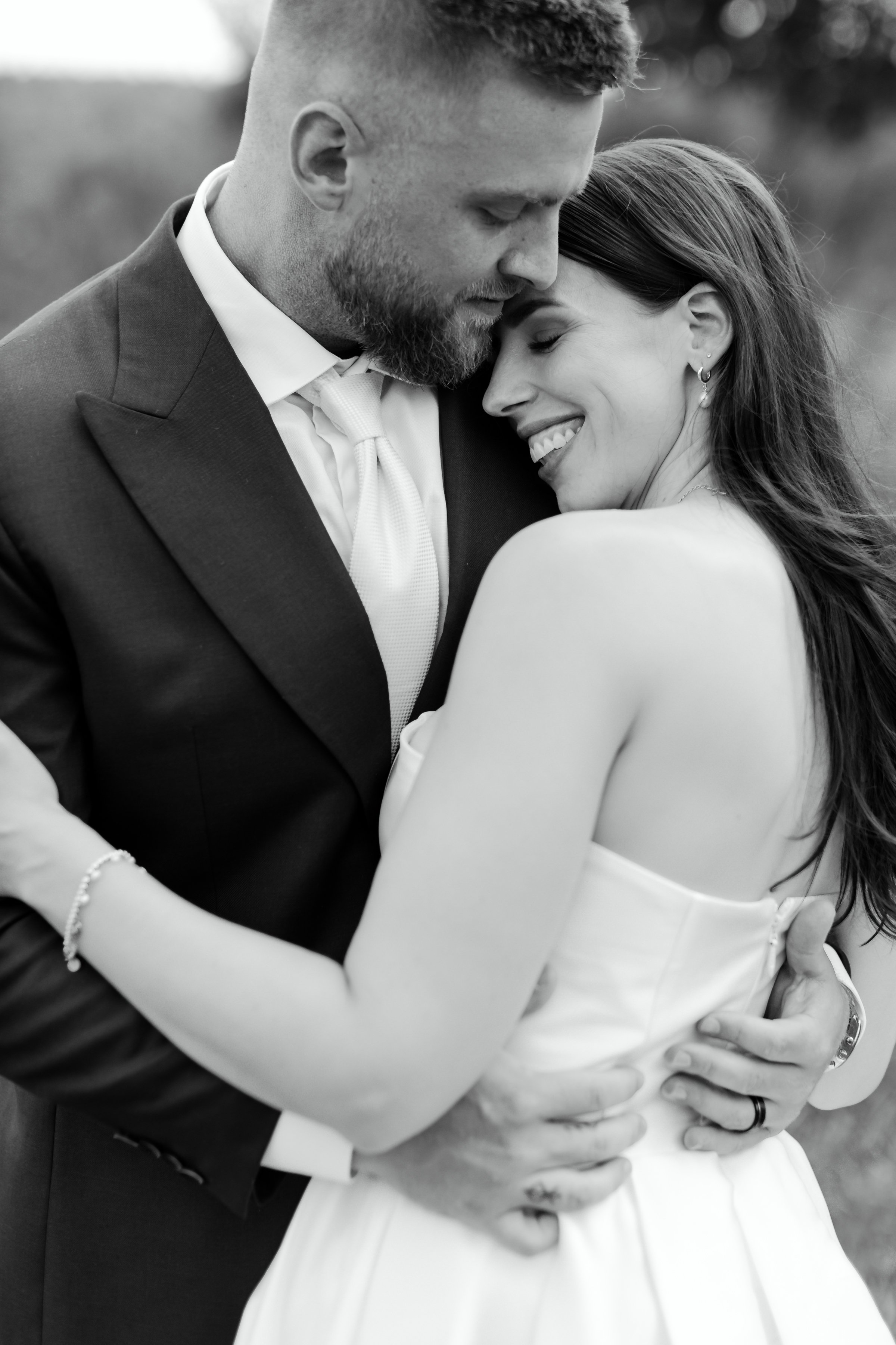 A black-and-white photo of a couple embracing, with their faces close together and the woman smiling with her eyes closed. The man has a beard and short hair, wearing a suit, while the woman has long hair and wears a strapless dress and earrings.