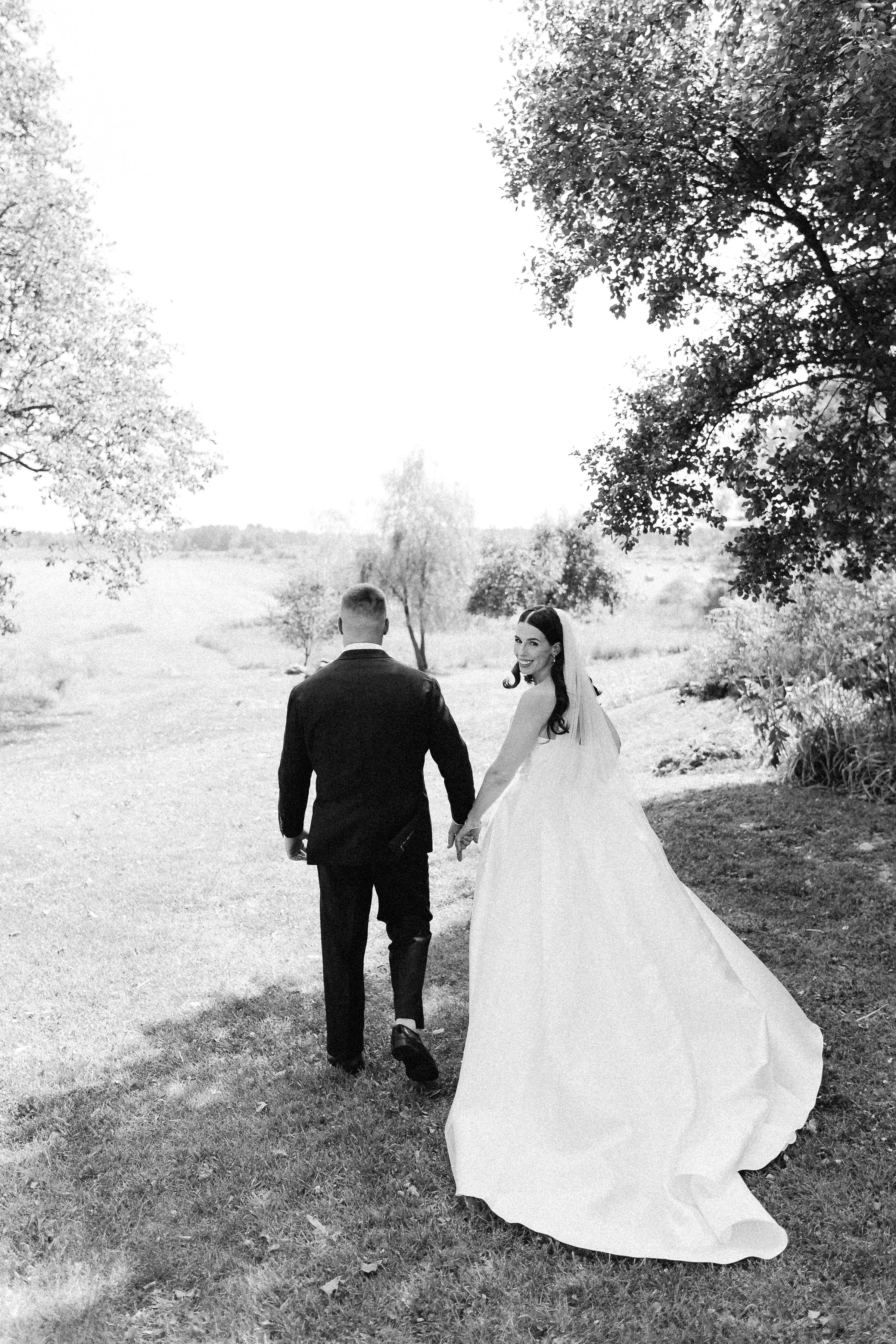 Black and white photo of a wedding couple walking hand in hand outdoors, with trees and a grassy landscape in the background.