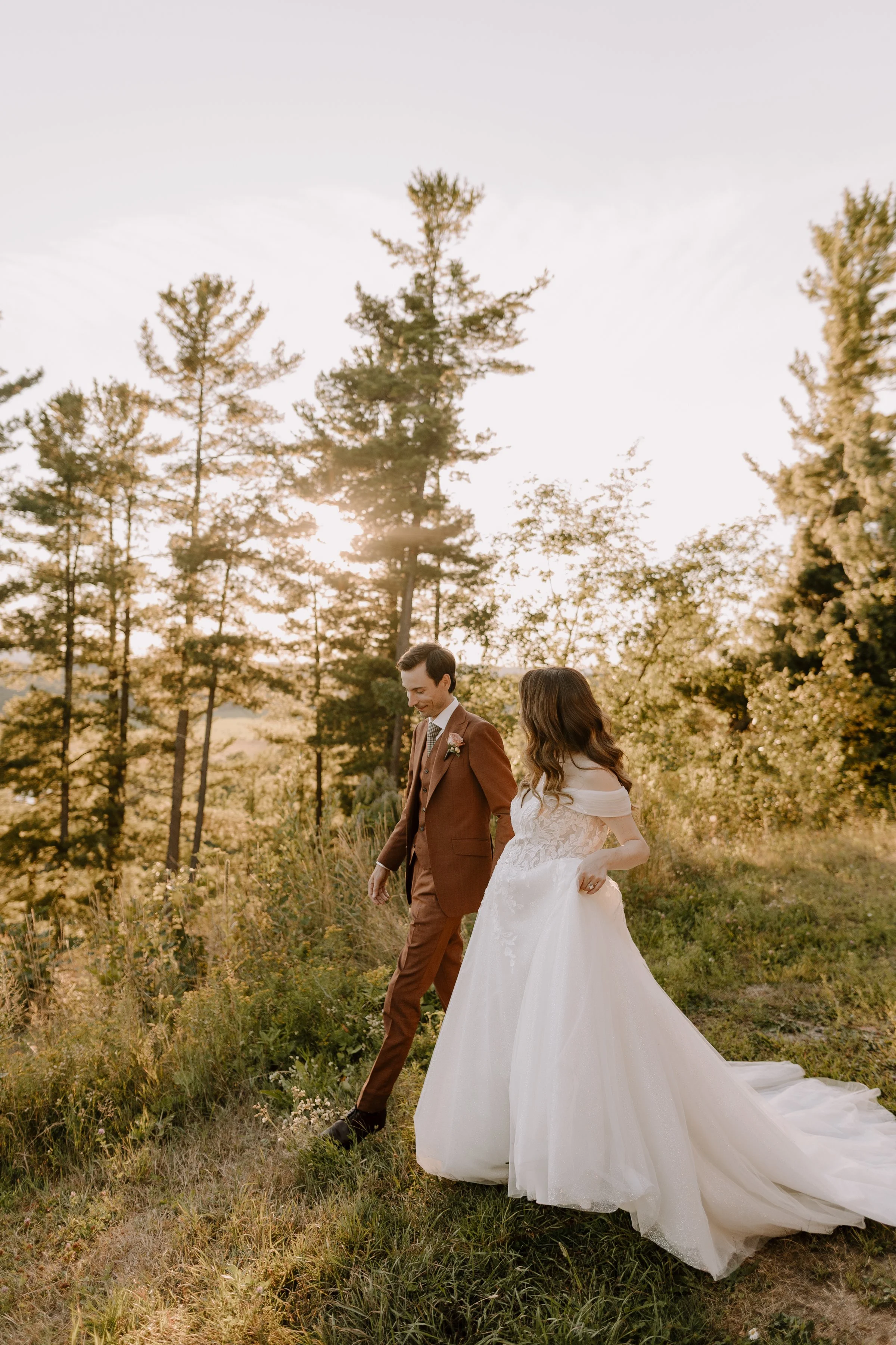 A bride and groom walking hand-in-hand through a grassy field at sunset, surrounded by trees.