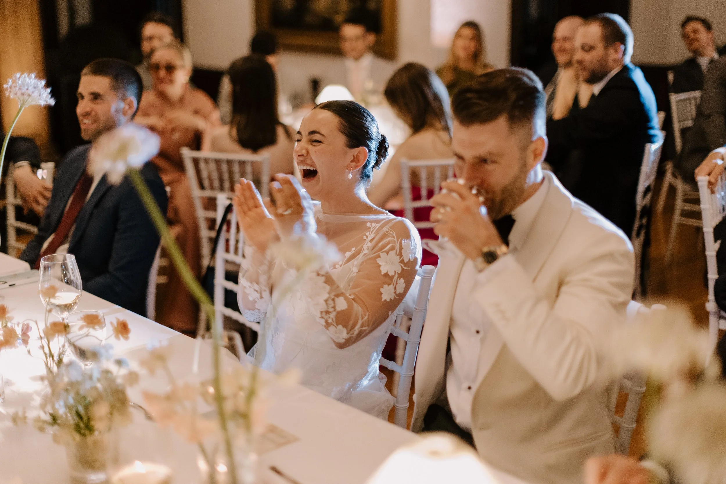A woman in a white wedding dress laughing and clapping at a wedding reception, seated at a table with floral decorations, surrounded by guests in formal attire.