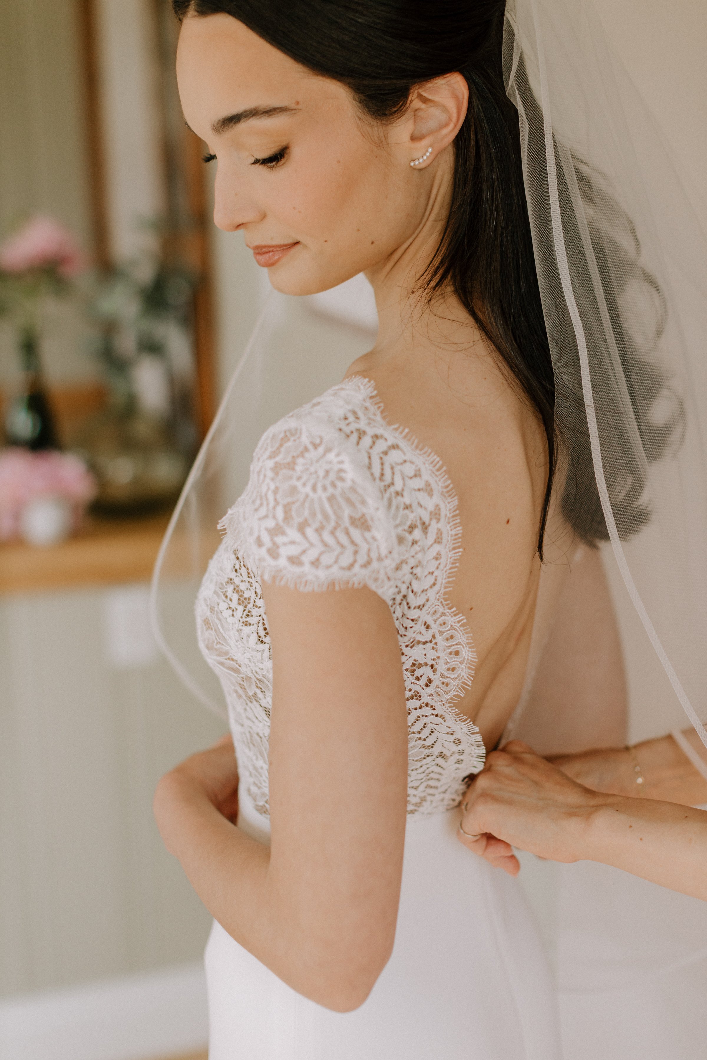 A bride in a white lace wedding dress and veil with a pearl earring, standing indoors with someone behind her, with blurred flowers in the background.