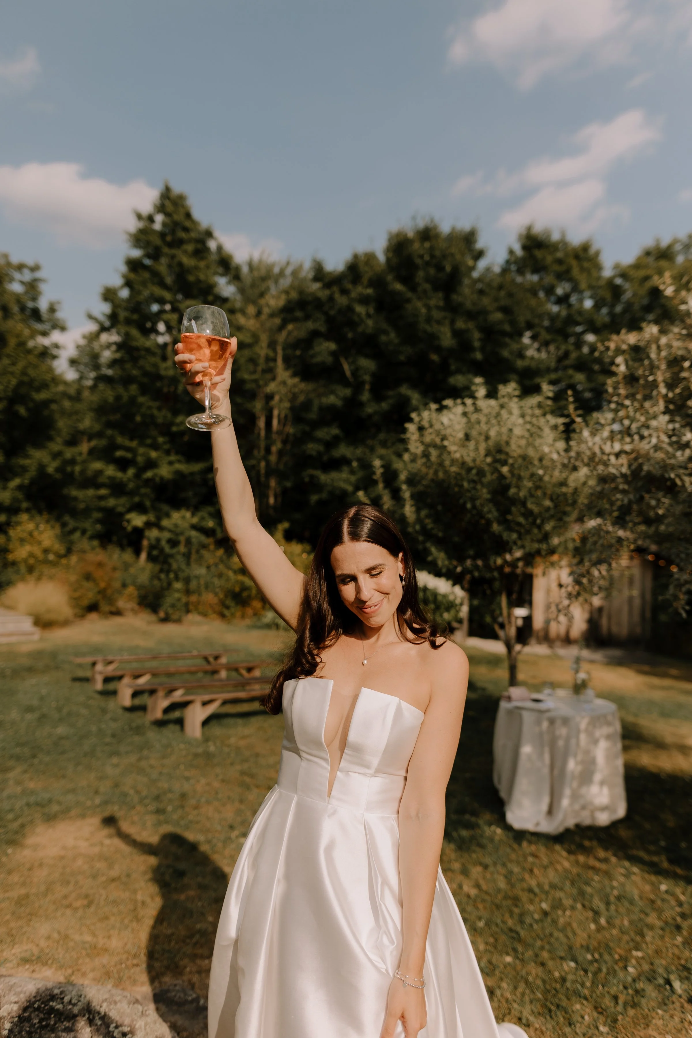 A woman in a white strapless wedding dress holding a glass of rosé wine up in an outdoor park setting.