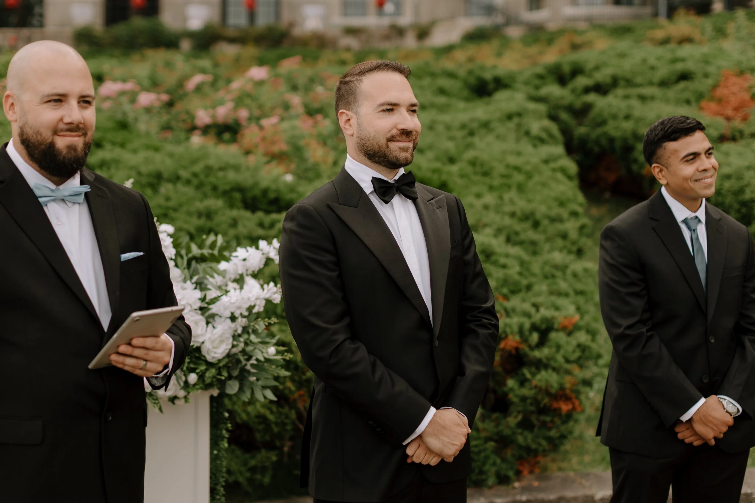 Three men in tuxedos stand outdoors during a wedding ceremony, with greenery and flowers in the background.