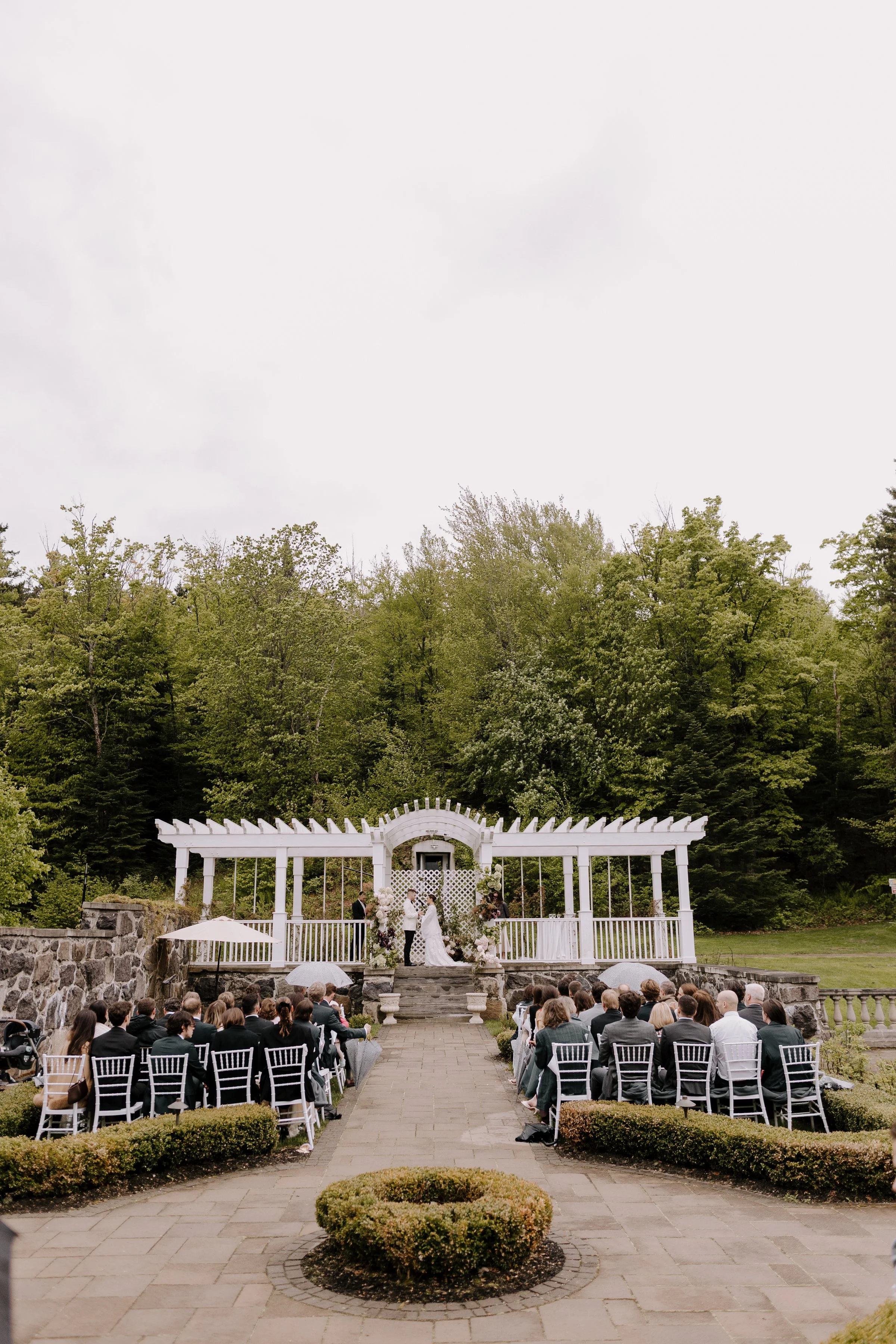 A wedding ceremony taking place outdoors on a cloudy day at a white pergola with a bride and groom standing in front of an officiant, surrounded by guests seated in white chairs, with lush green trees in the background.