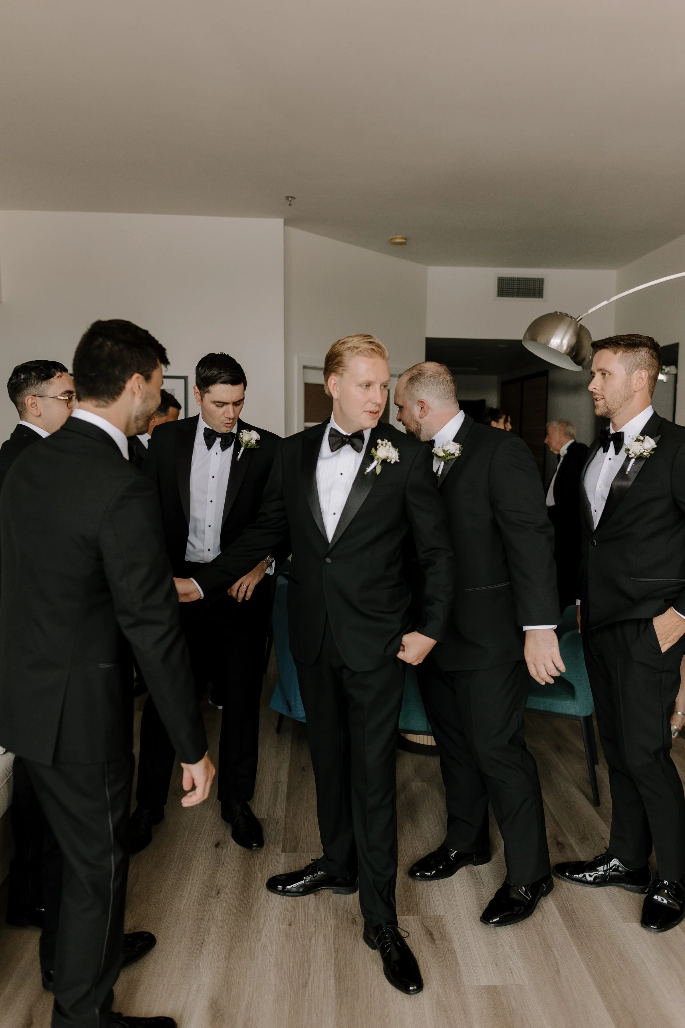 A group of men dressed in black tuxedos with white shirts and black bow ties, standing in a circle, holding hands, in an indoor setting.