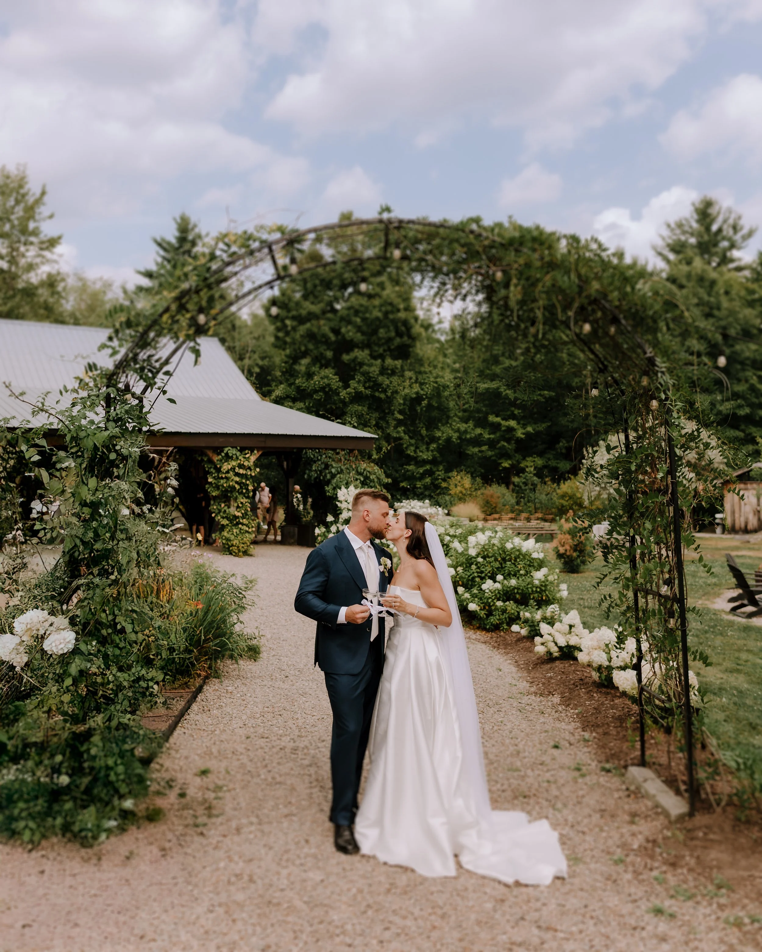A bride and groom sharing a kiss at their outdoor wedding, surrounded by greenery and white flowers, with a cloudy sky overhead.