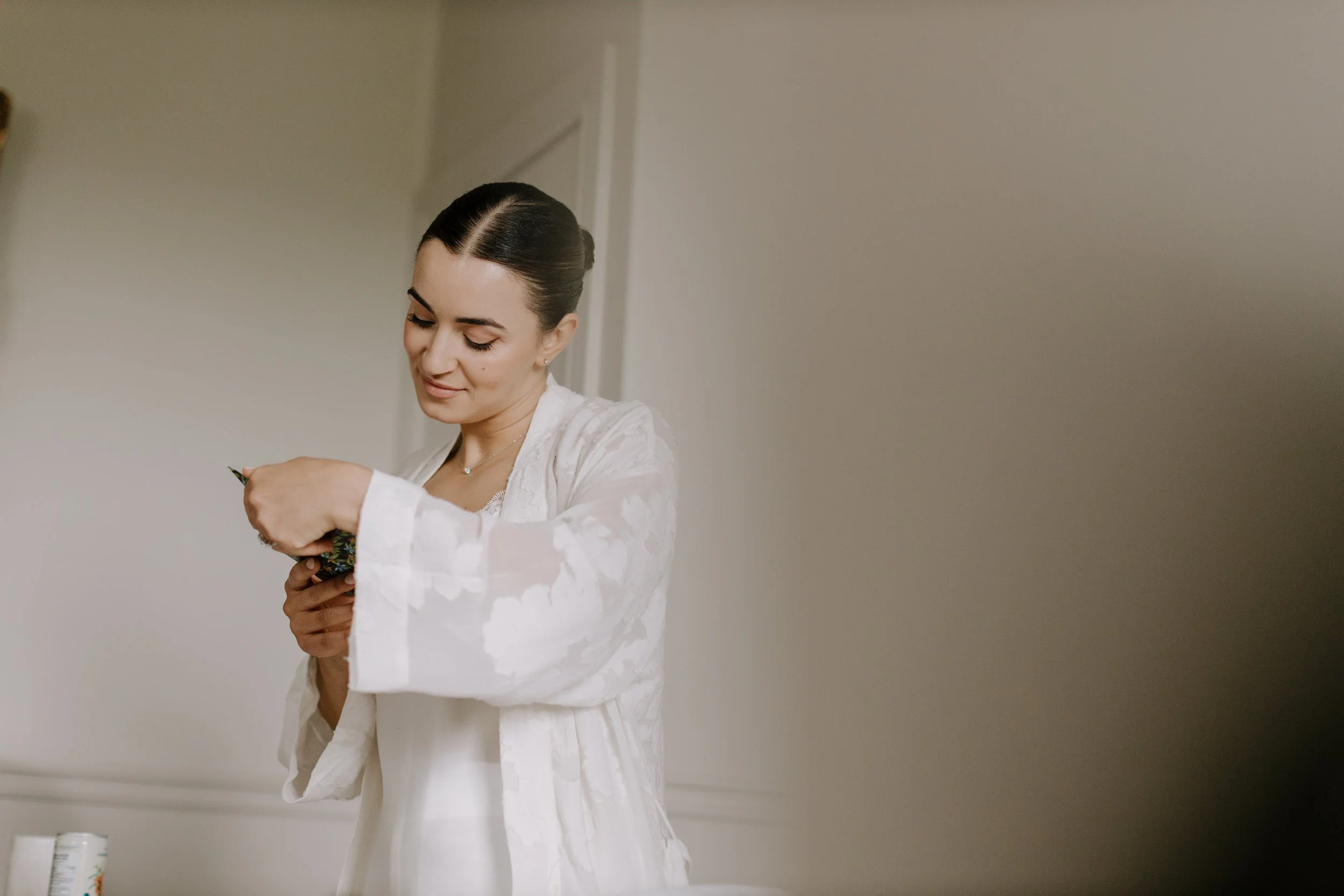 A woman with dark hair styled in an elegant bun, wearing a white robe, standing against a plain wall, smiling slightly while looking down at her hands, which are holding a small object.
