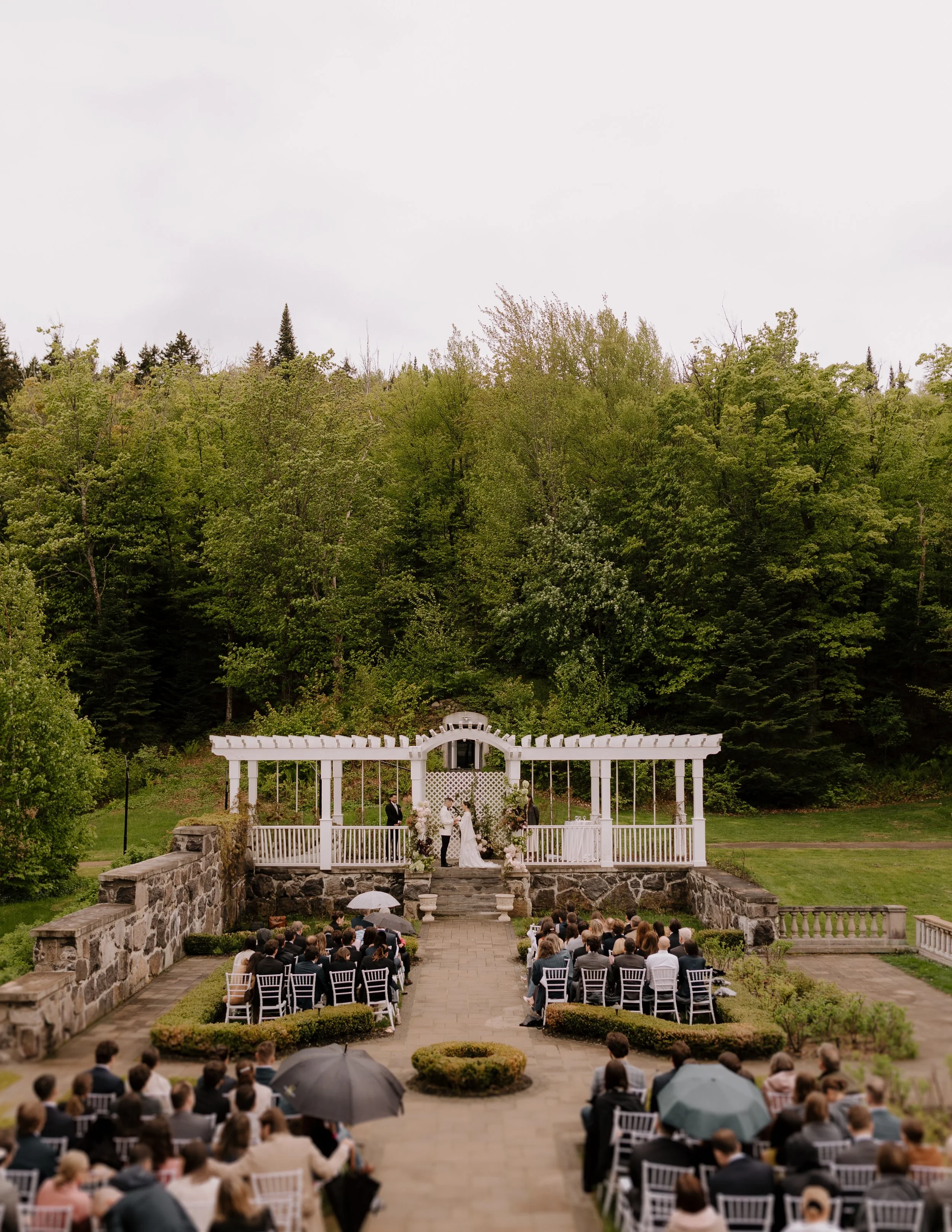 Outdoor wedding ceremony taking place on a stone-paved terrace with a white gazebo and floral decorations, surrounded by green trees, with guests seated in white chairs under umbrellas on a cloudy day.