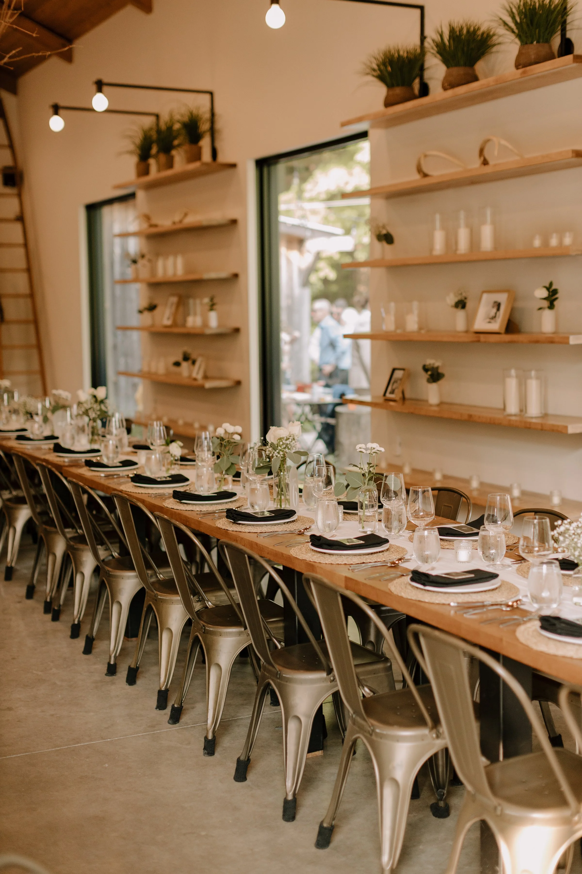 A long wooden table set for a meal with black napkins, clear water glasses, and small flower arrangements. The room has large windows, white walls, wooden shelves with decorative items, and green indoor plants on top. The chairs are metallic with bla