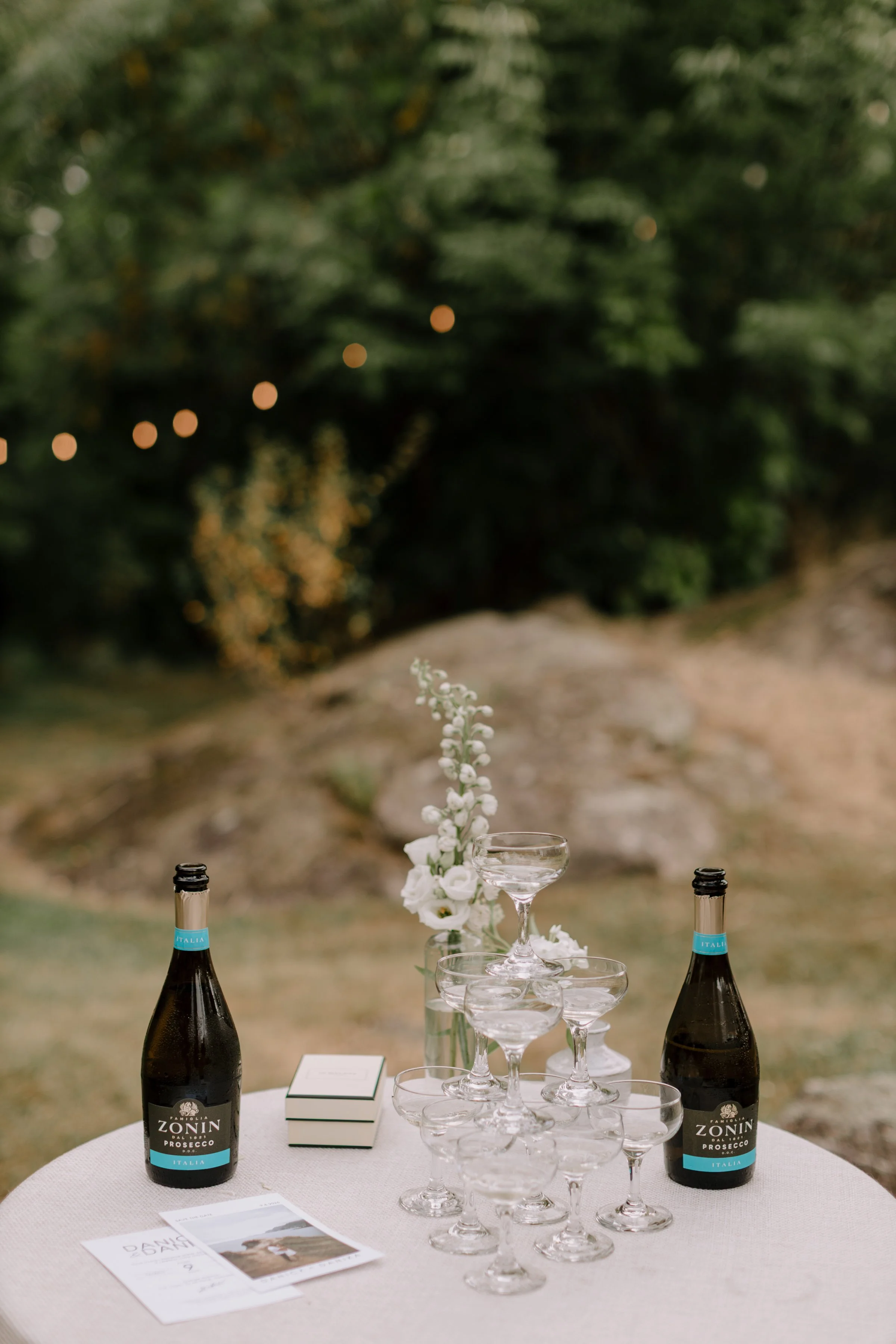 An outdoor table setup with two bottles of Zonin Prosecco, a champagne glass, a flower arrangement, and a small menu card, with a blurred green tree background.