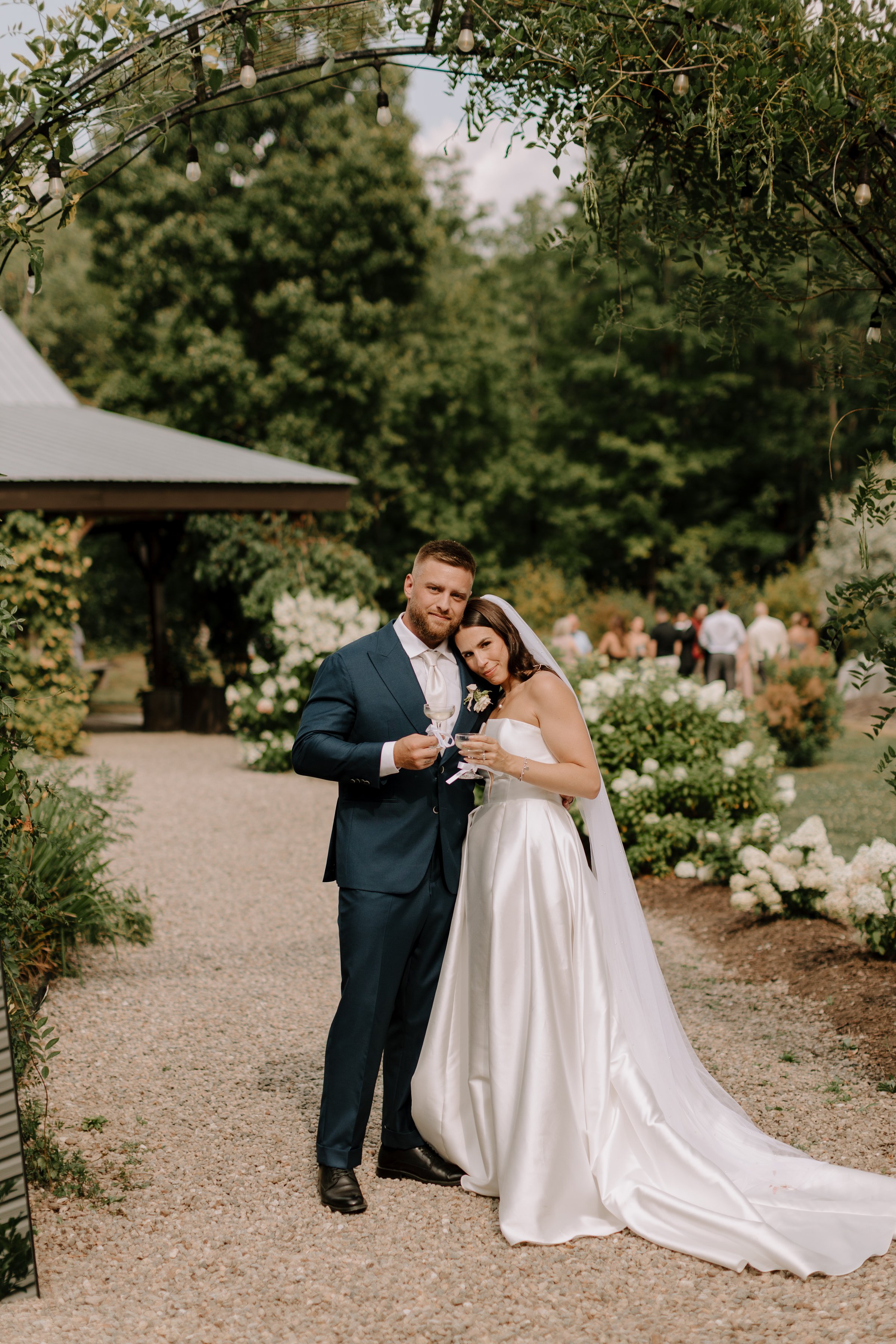 A newlywed couple in wedding attire posing outdoors at a garden wedding. The groom is wearing a navy suit and the bride is in a white strapless wedding gown with a train. They are holding glasses, possibly for a toast, and are standing close together