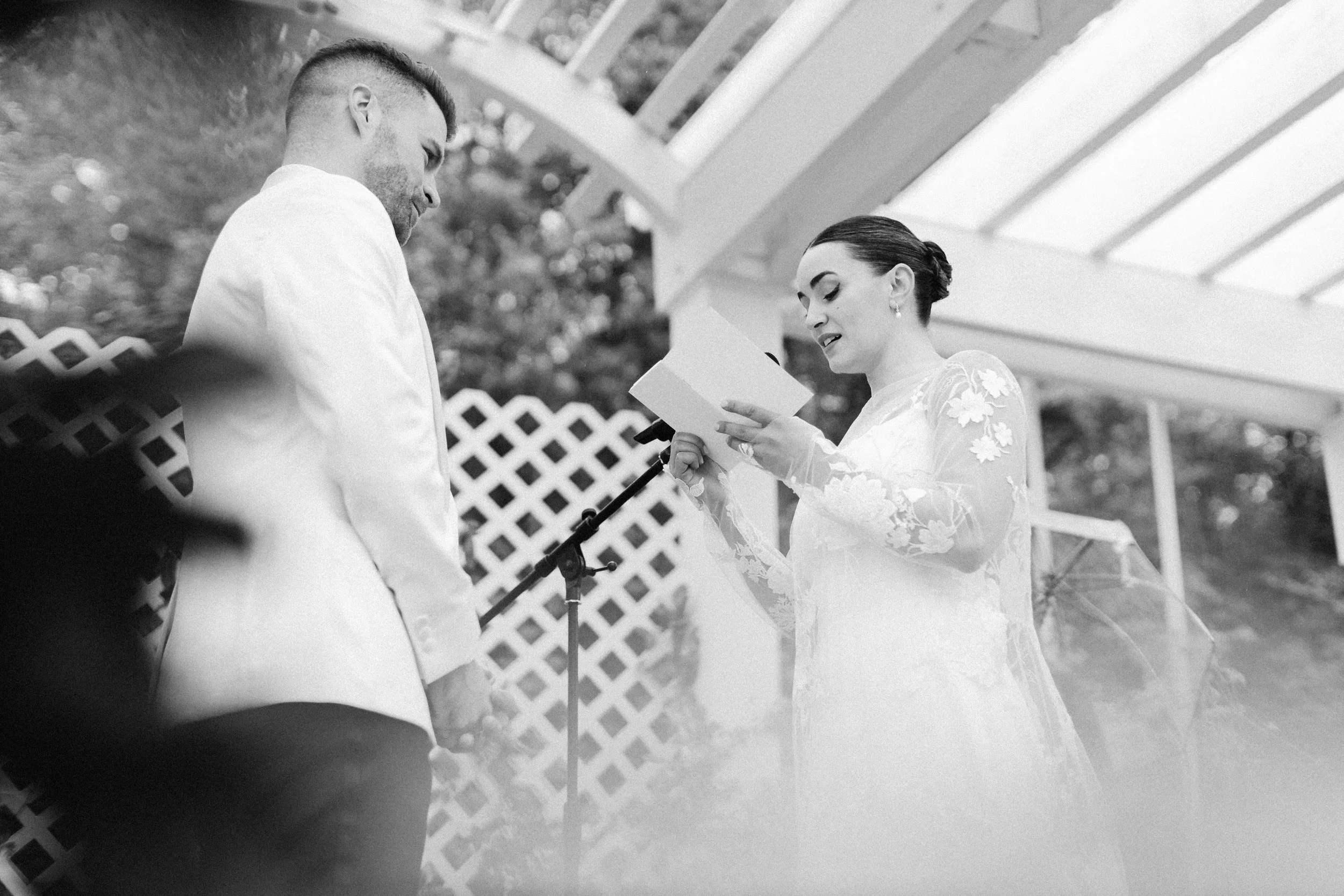A black and white photograph of a wedding ceremony, showing a woman reading from a booklet and a man standing opposite her, outdoors under a gazebo with a lattice fence in the background.