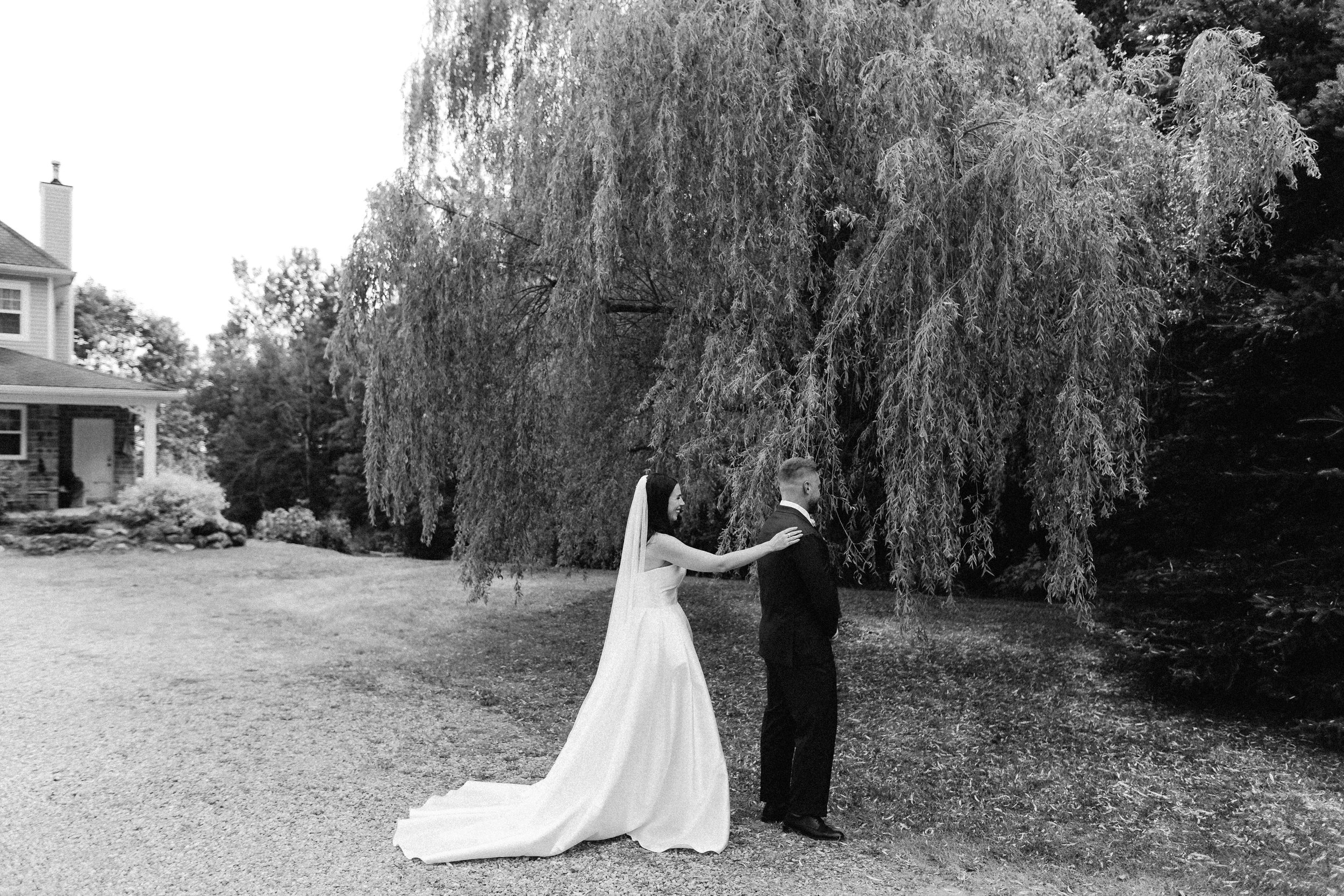A bride and groom standing outdoors beneath a large, leafy tree, with the bride reaching out to touch the groom's shoulder, during their wedding day.