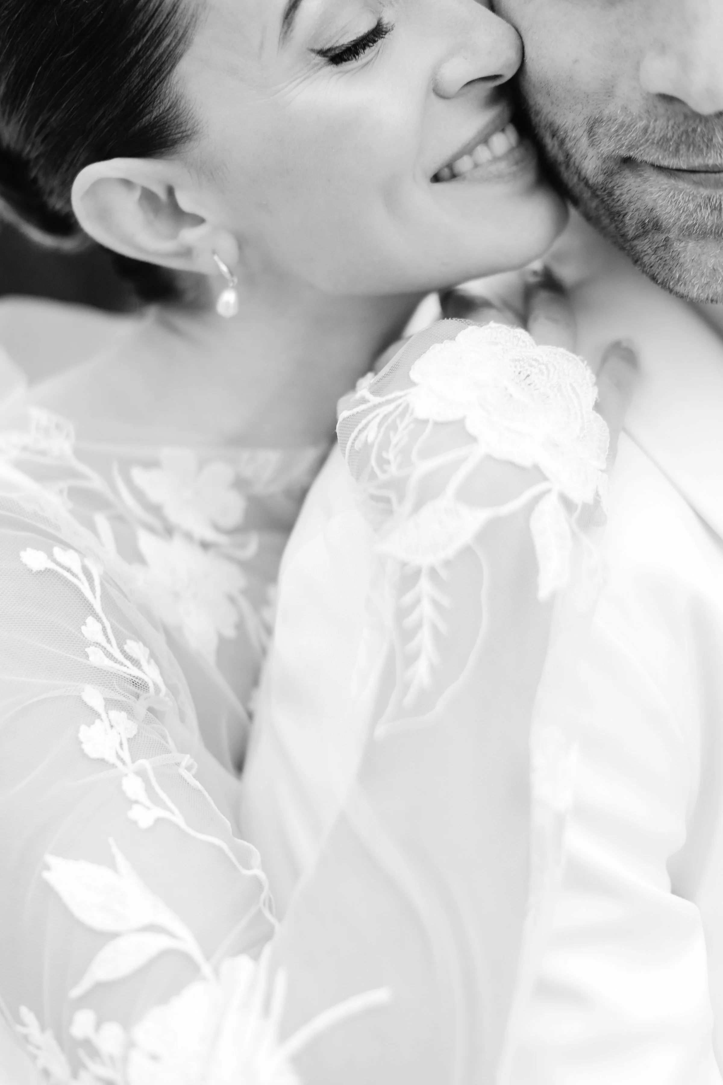 A close-up black and white photograph of a couple sharing a tender moment, with the woman smiling and the man’s face partially visible, both dressed in elegant attire.