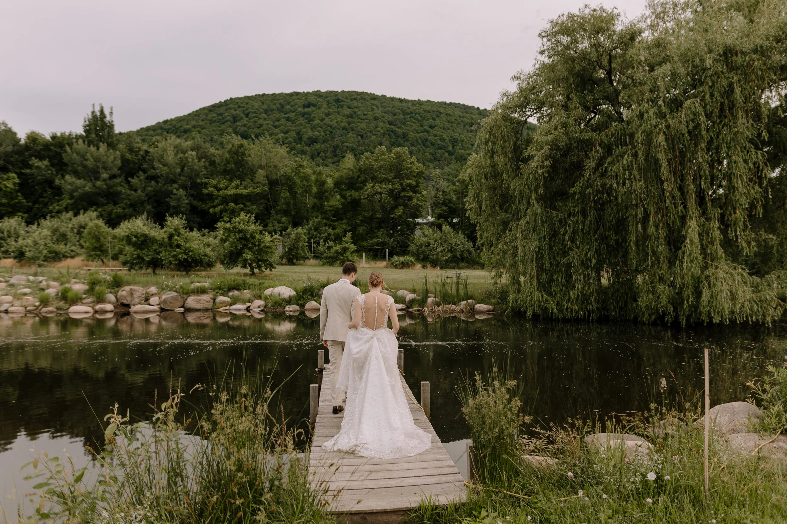 A bride and groom standing on a dock by a lake with lush green trees and a hill in the background, during daylight.