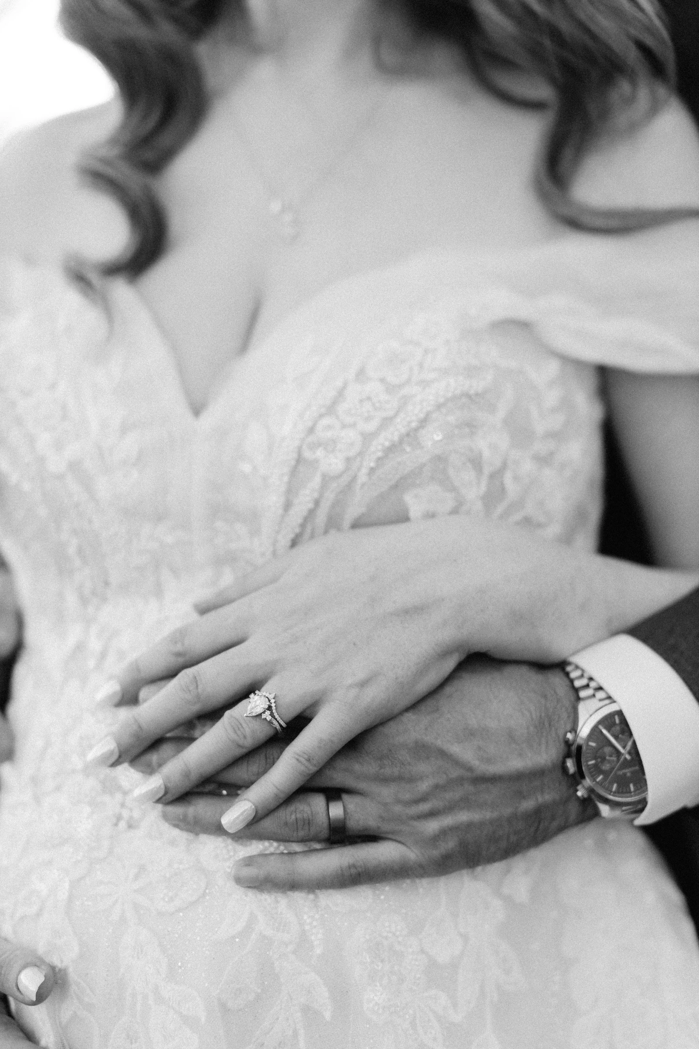 A close-up of a couple's hands intertwined, showing wedding rings and a wristwatch, with a lace wedding dress in the background.