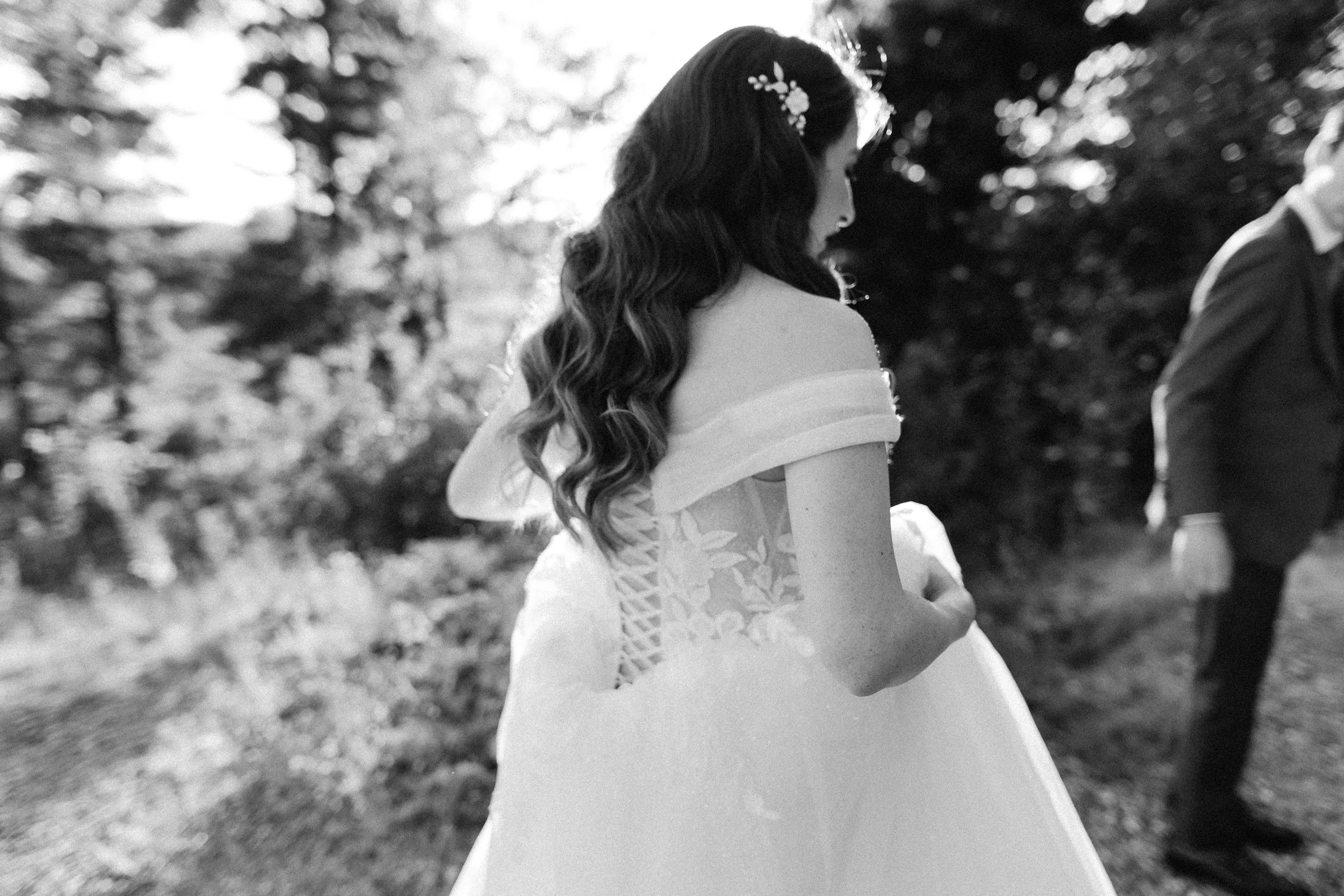 Close-up of a woman in a wedding dress with long, wavy hair, wearing glasses and floral hair accessories, standing outdoors with trees in the background, during daytime.