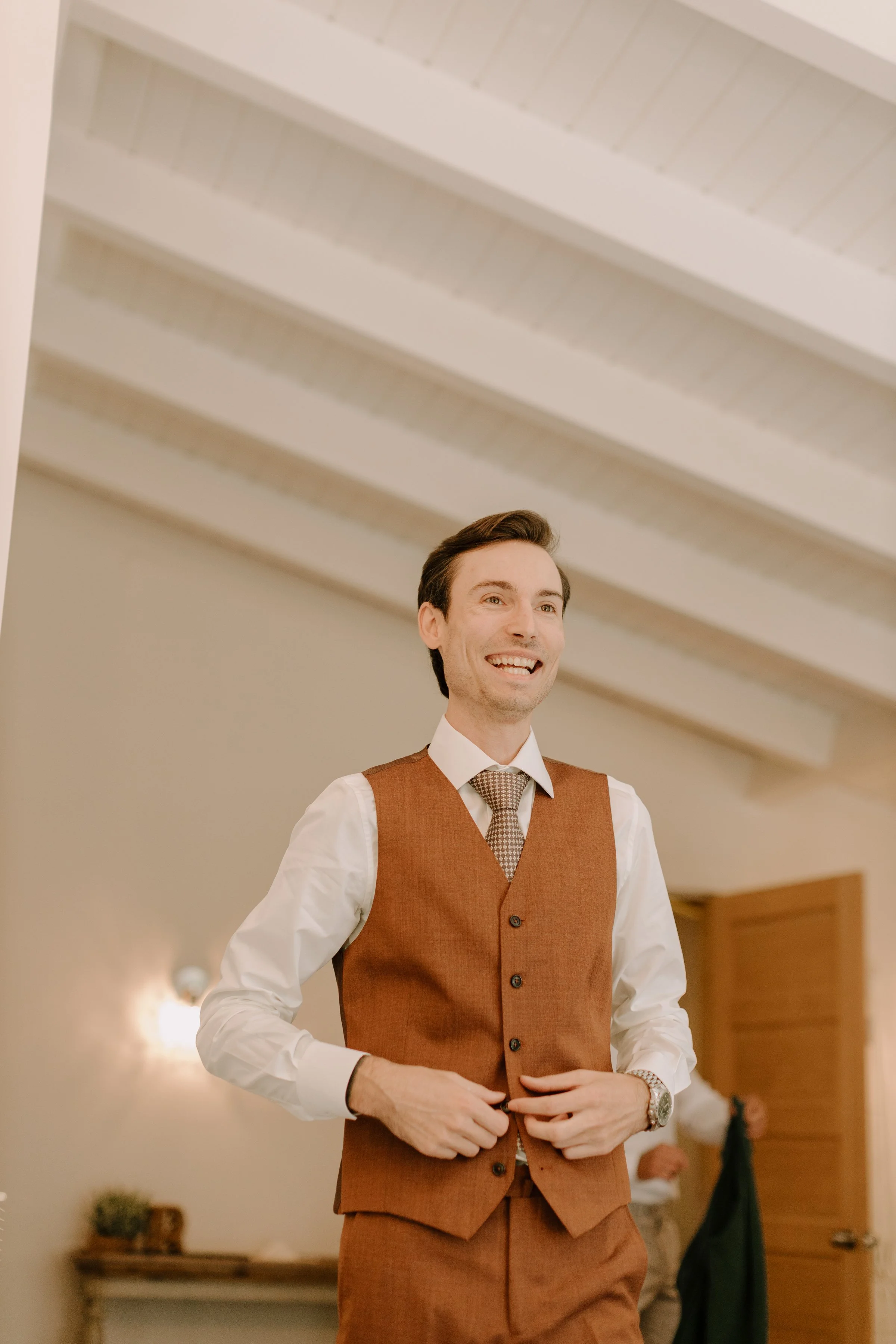 A man smiling and dressed in a white shirt, brown vest, and matching brown pants, standing inside a room with warm lighting, a wooden door, and a small table with plants in the background.