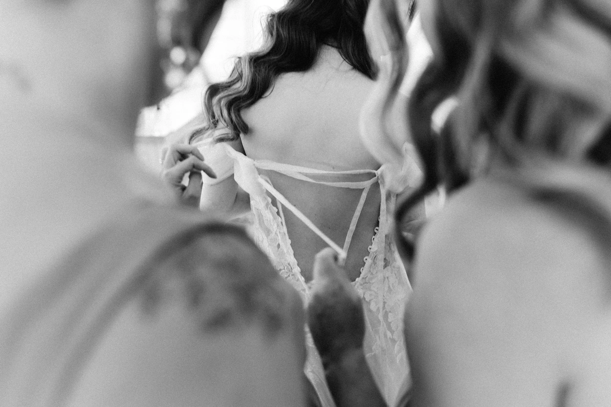 Black and white photo of a person, possibly a bride, adjusting her wedding dress with lace details, seen from behind.
