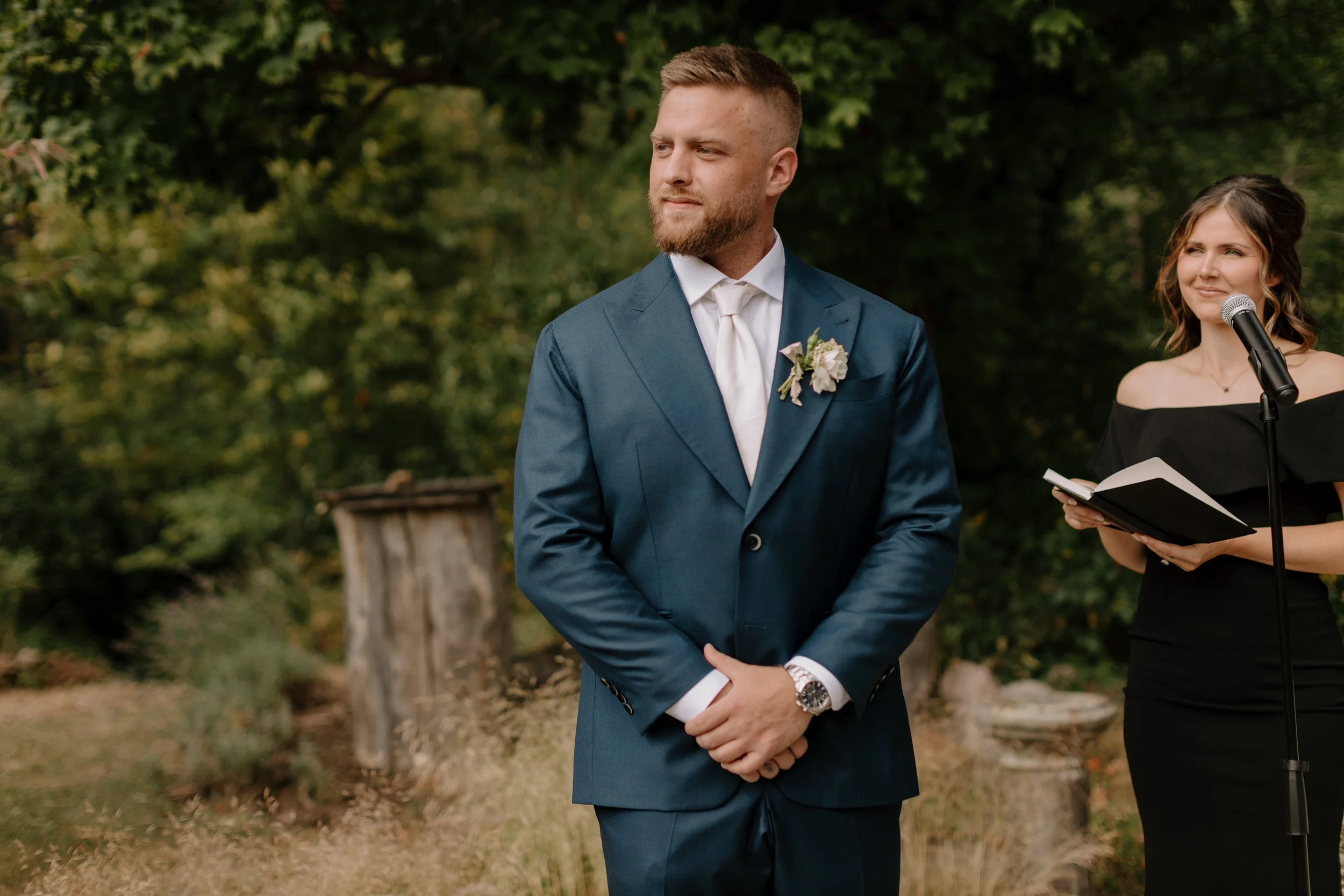 A man dressed in a blue suit and white shirt with a boutonniere, standing outdoors with trees in the background, during a formal event or wedding ceremony.