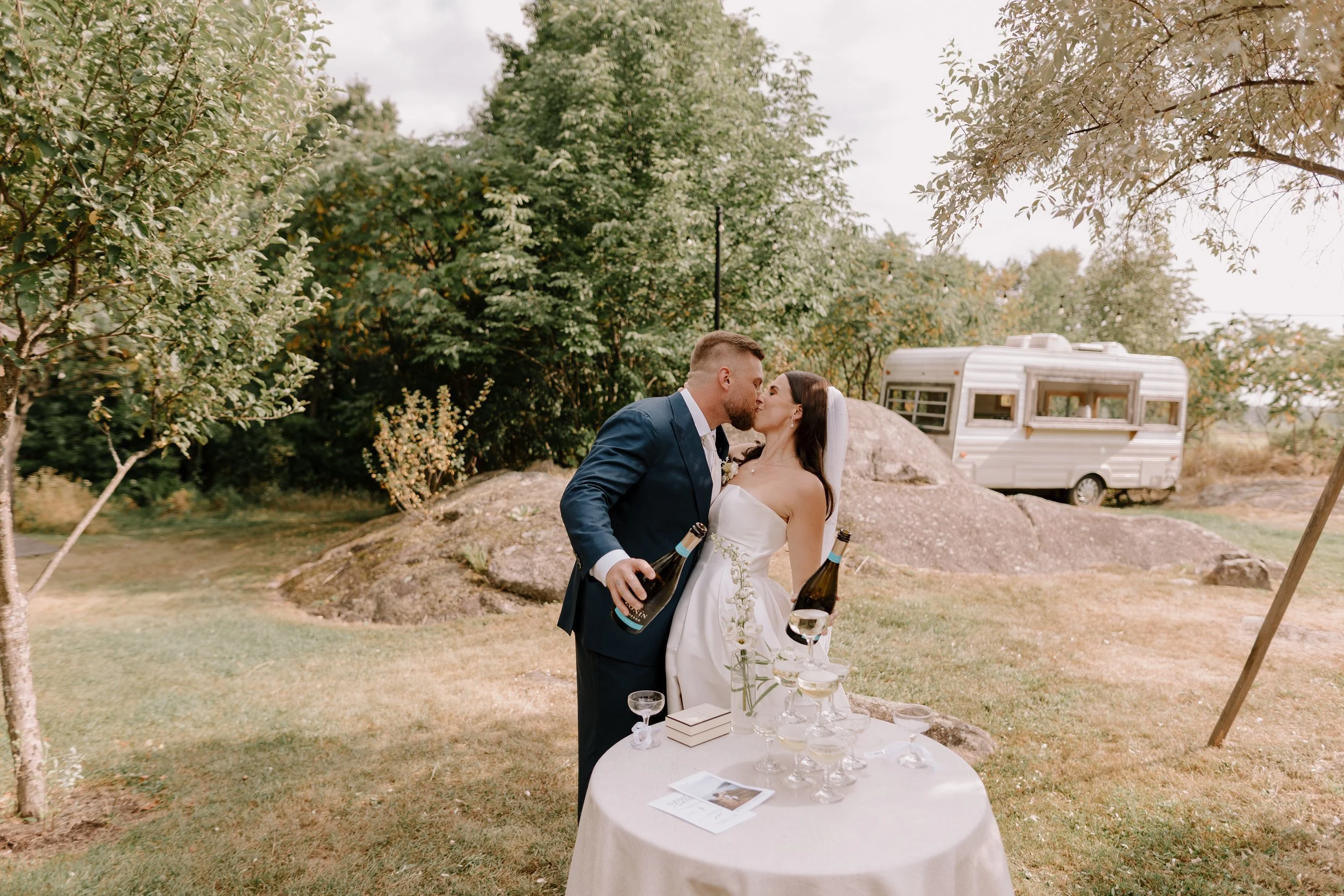 A bride and groom kissing outdoors during their wedding celebration, with a table of champagne glasses and wedding programs in the foreground and a vintage trailer in the background.