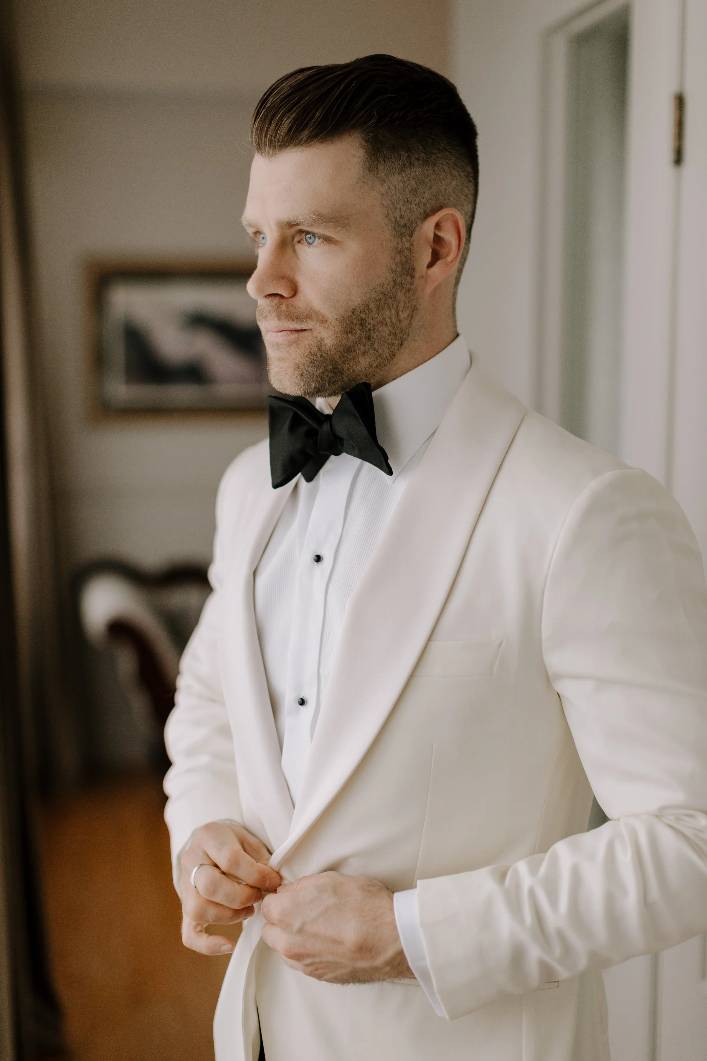 A man dressed in a white tuxedo with a black bow tie, adjusting his jacket. He has short, dark hair, blue eyes, and light facial hair. The background appears to be an indoor setting with framed artwork on the wall.