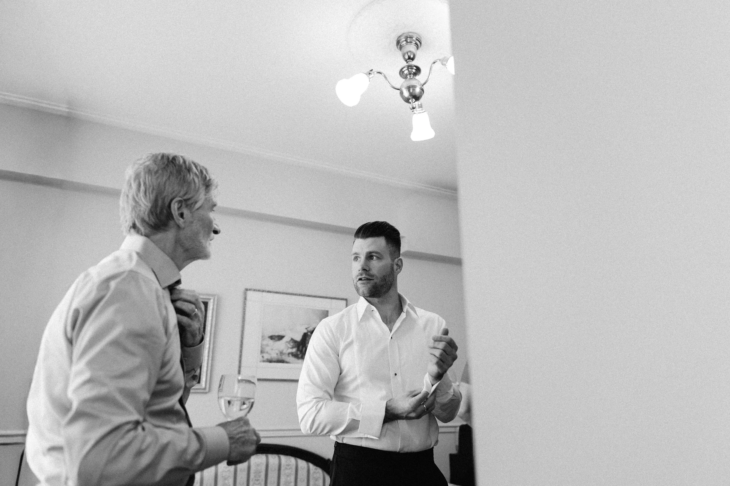 Two men in white shirts, one older with light-colored hair holding a glass of wine, and one younger with dark hair, engaged in conversation in a room with framed artwork on the wall.