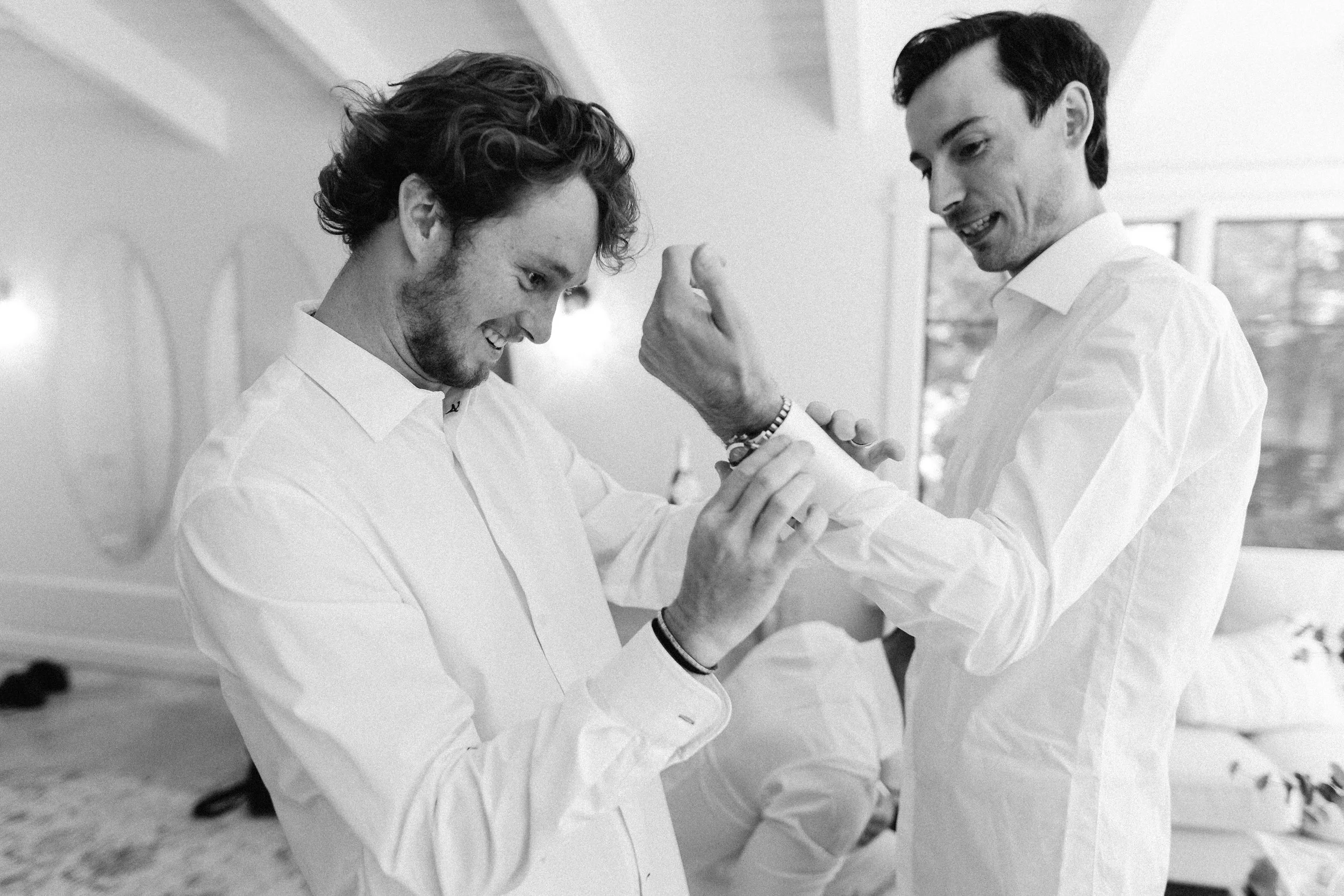 Two men in white dress shirts smiling and playfully engaging with each other, one adjusting the other's sleeve, indoors with large windows in the background.