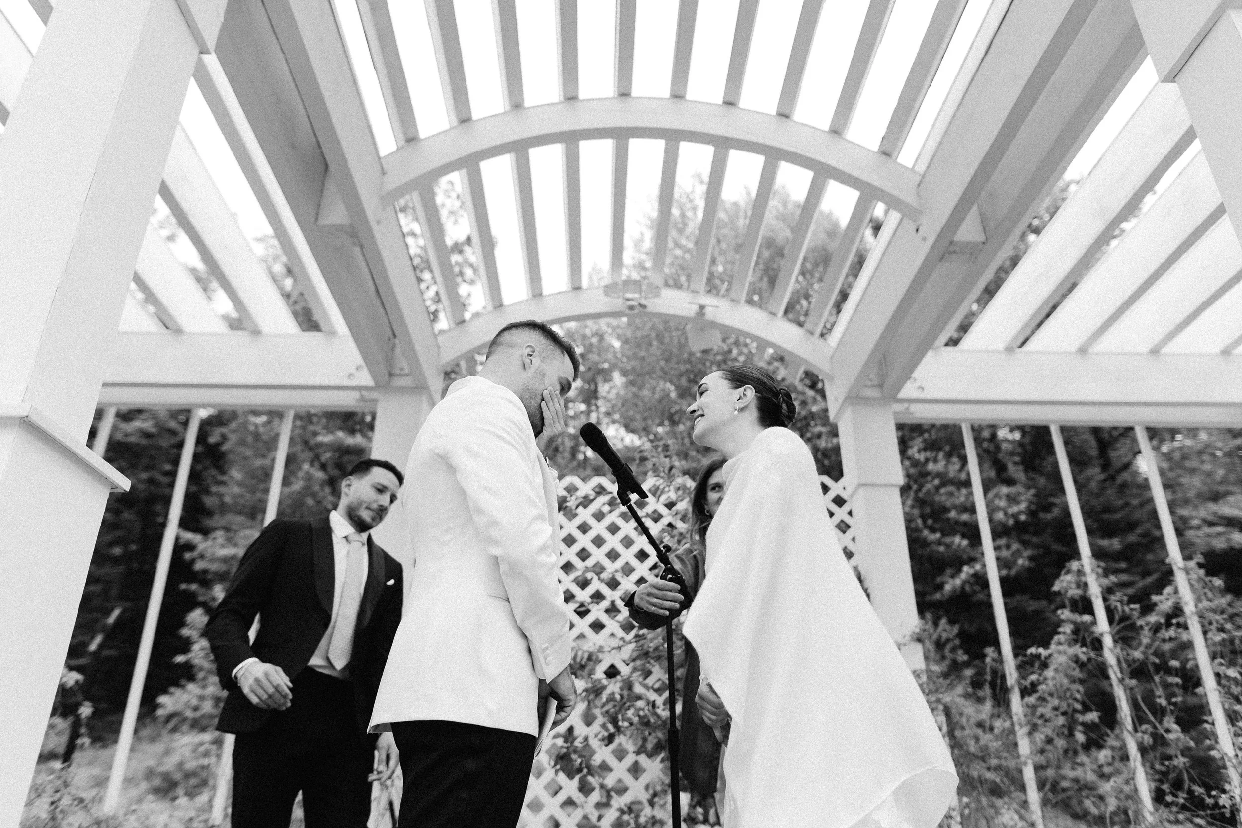 A black and white photo of a wedding ceremony with a bride and groom exchanging vows under a white wooden arch. The bride holds a microphone, and the groom has his hand over his face, possibly crying.