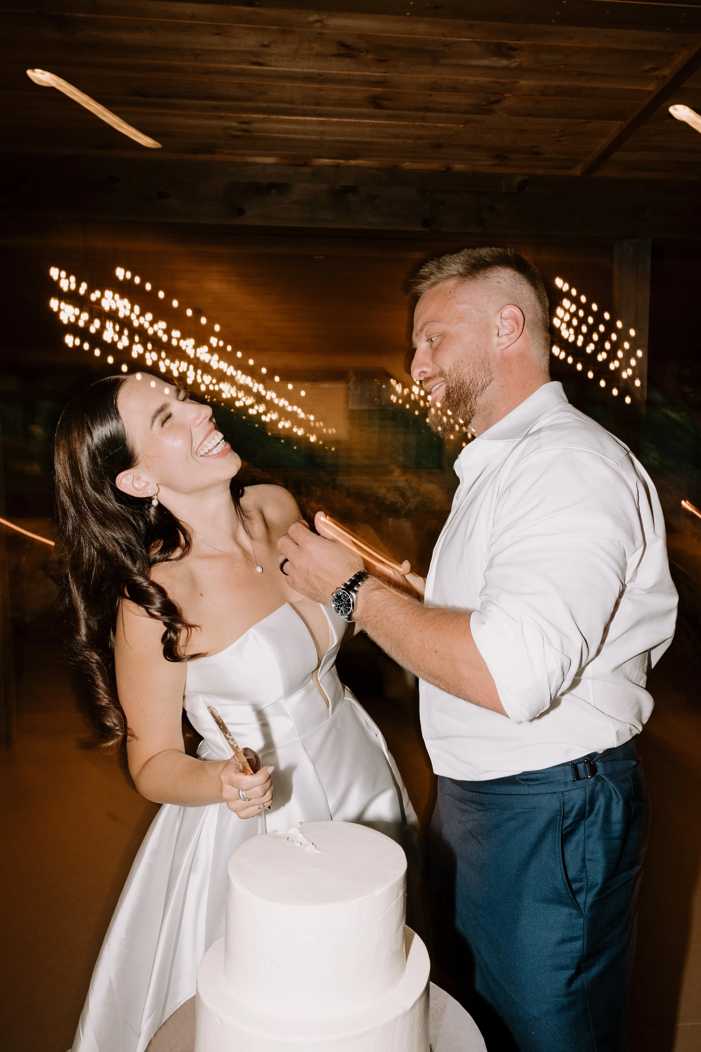 A smiling woman in a white dress with dark hair, and a man with short hair and a beard in a white shirt, are at a wedding cake table with a round white cake. They are dancing or celebrating together in a dimly lit room with string lights overhead.