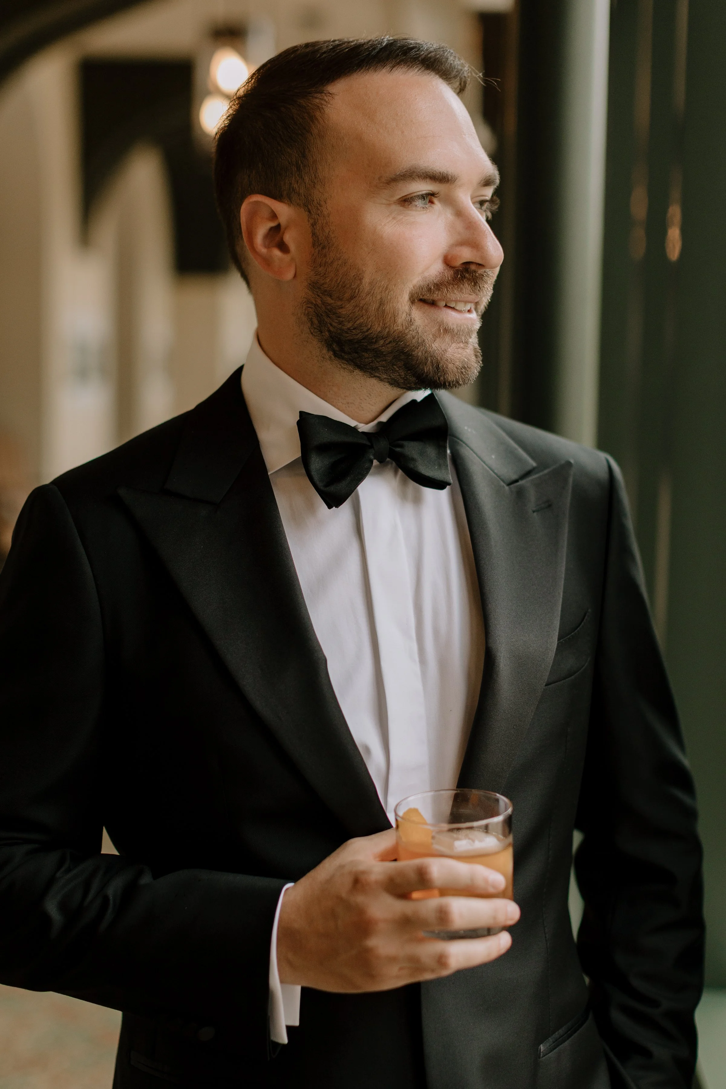 A man dressed in a tuxedo with a bow tie holding a cocktail glass, looking to the right and smiling, at an indoor event.