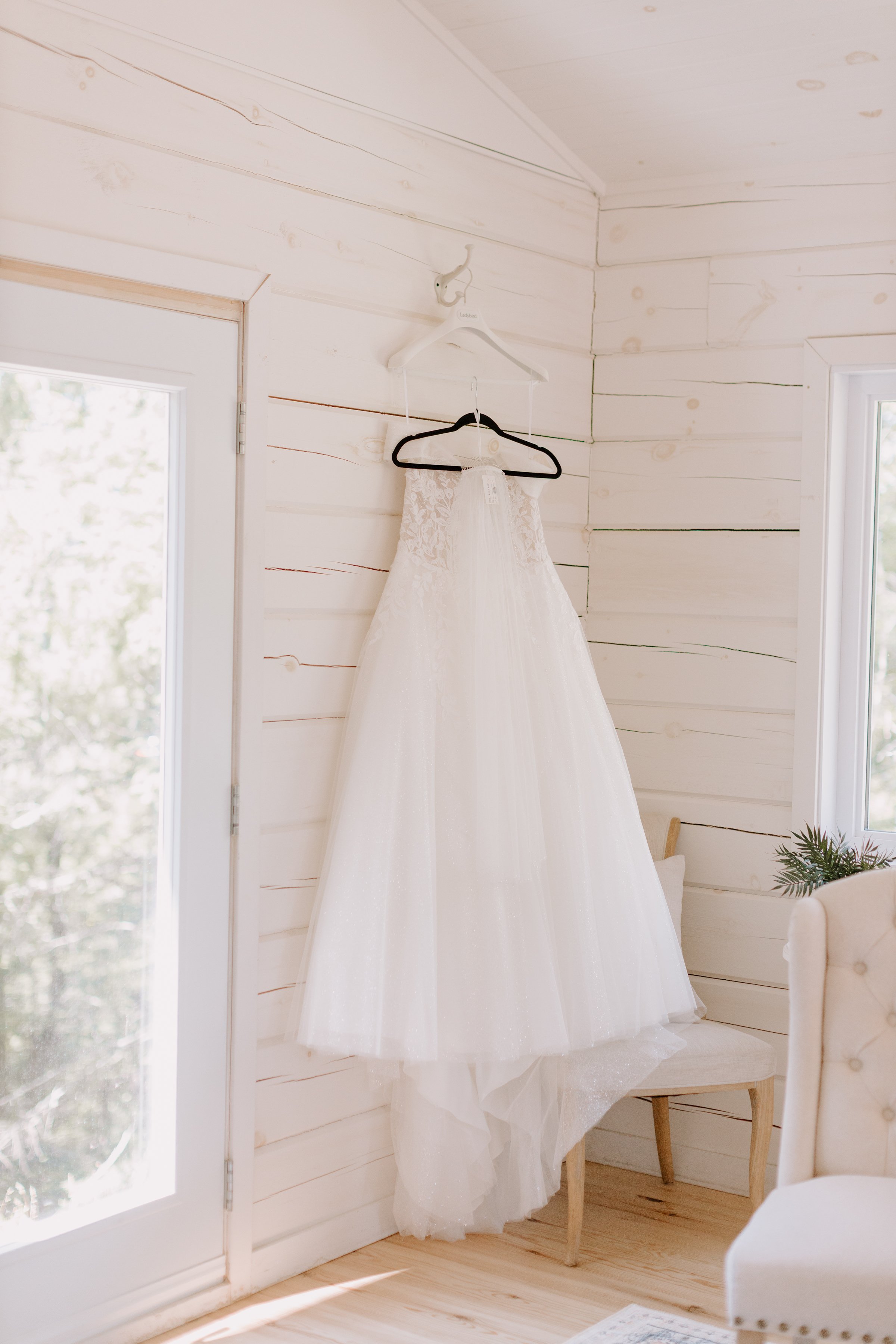 A white wedding dress hanging on a hanger in a bright, cozy room with light-colored wooden walls and floor, near a window with natural light shining in.