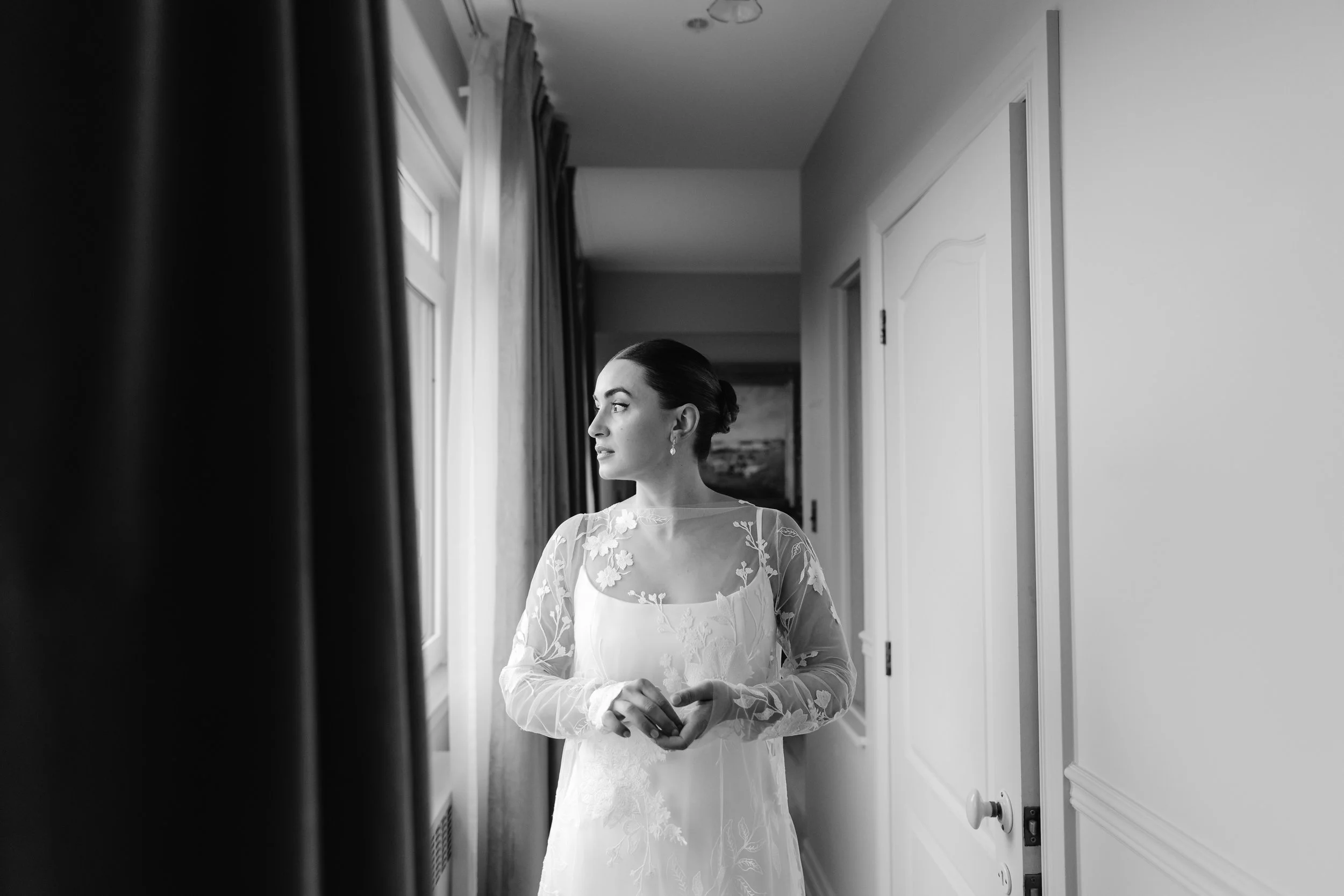 A woman in a lace wedding dress looking out a window in a bright, minimalistic room.