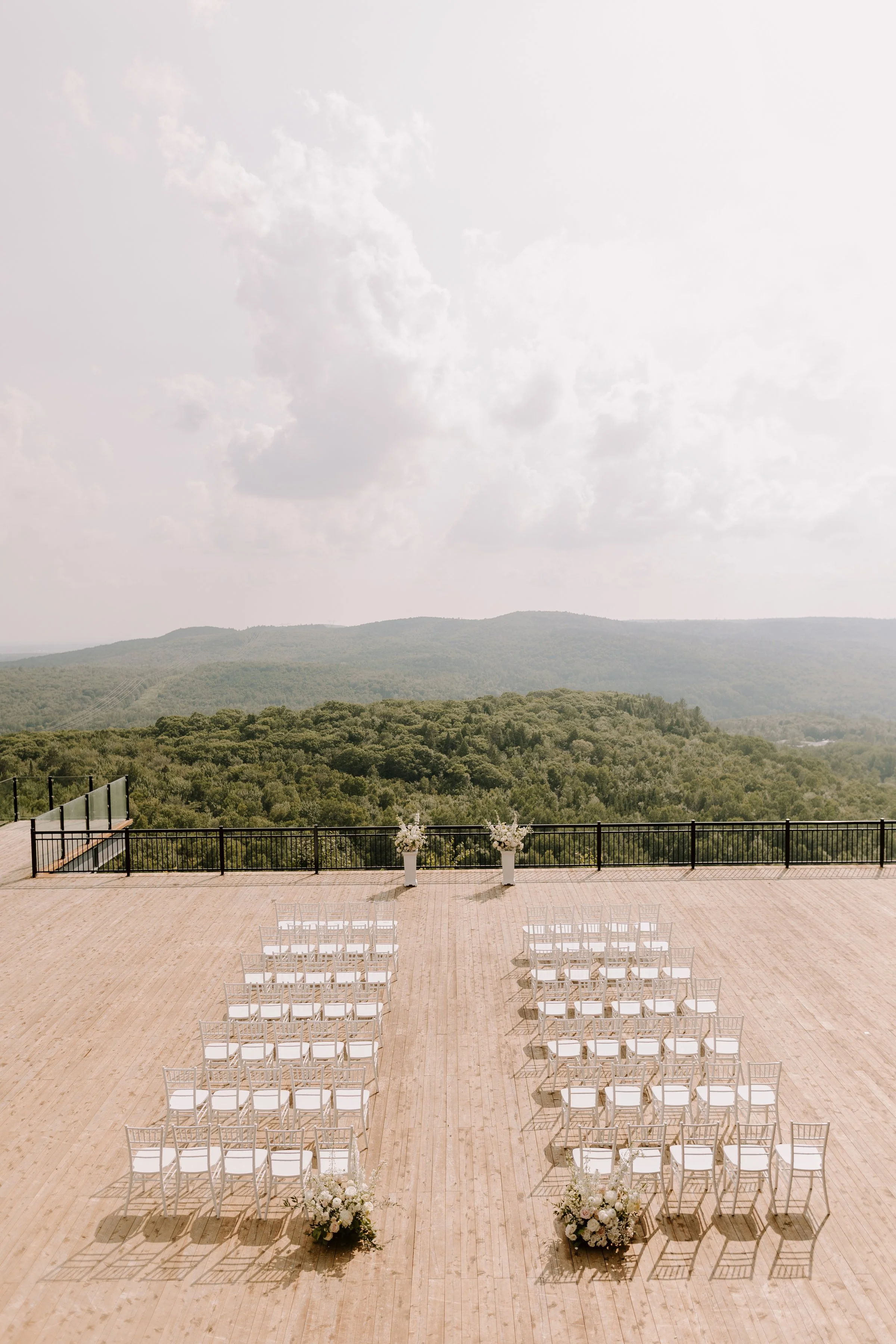 Outdoor wedding setup on a wooden deck with white chairs facing an altar with floral decorations. The deck overlooks a green forested landscape with distant hills and a partly cloudy sky.