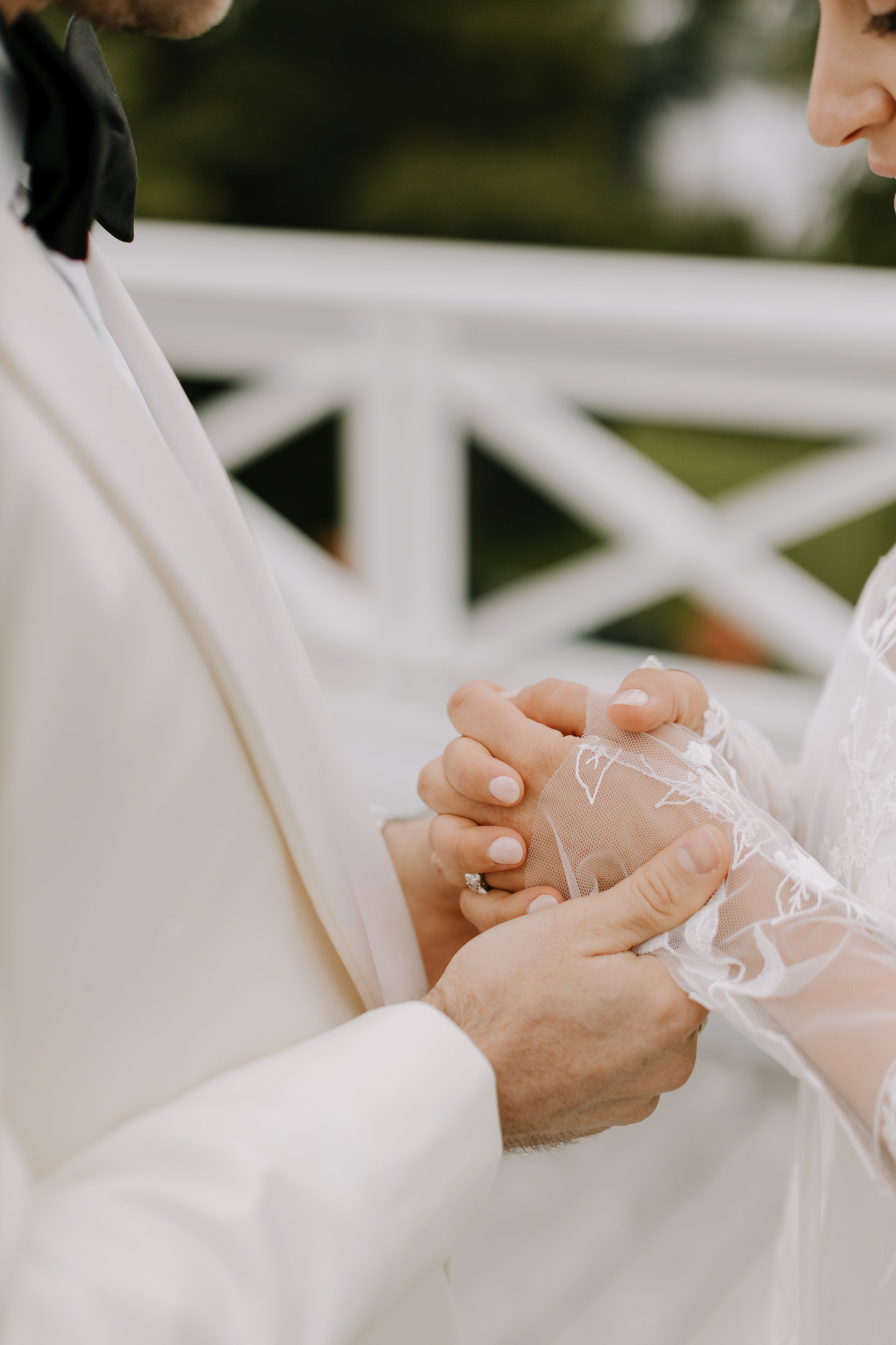 A bride and groom holding hands during their wedding ceremony, with a focus on their clasped hands and wedding attire. The bride’s hand is wearing a lace glove and an engagement ring. The groom is wearing a white suit and black bow tie.