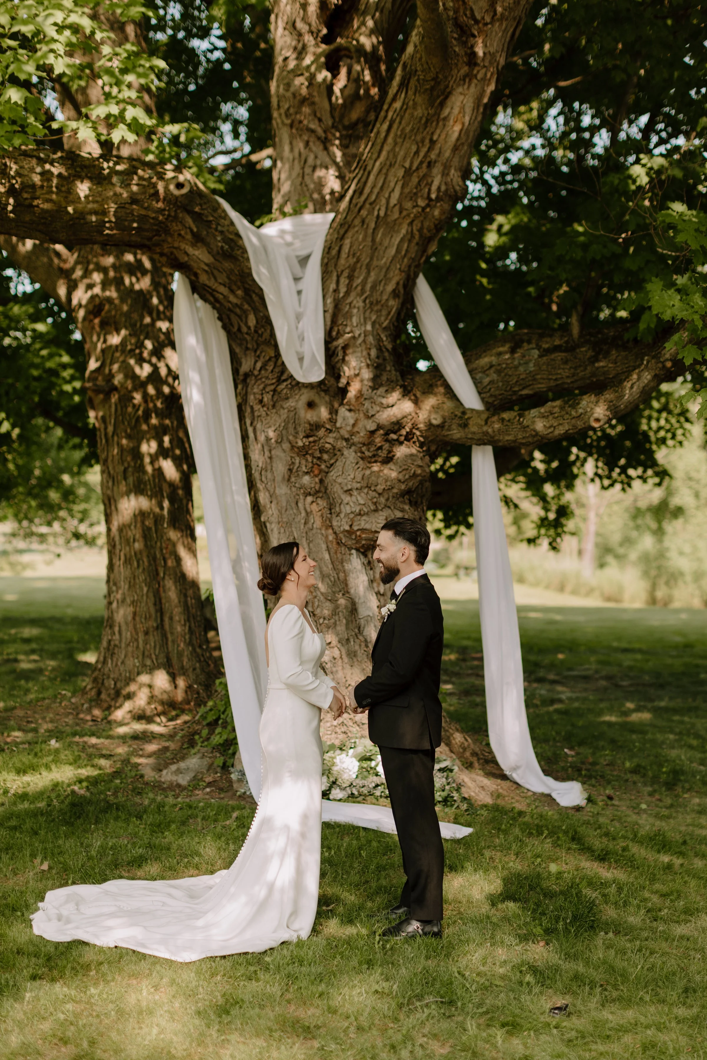 A couple getting married outdoors under a large tree with white drapes hanging from its branches, holding hands and looking at each other.