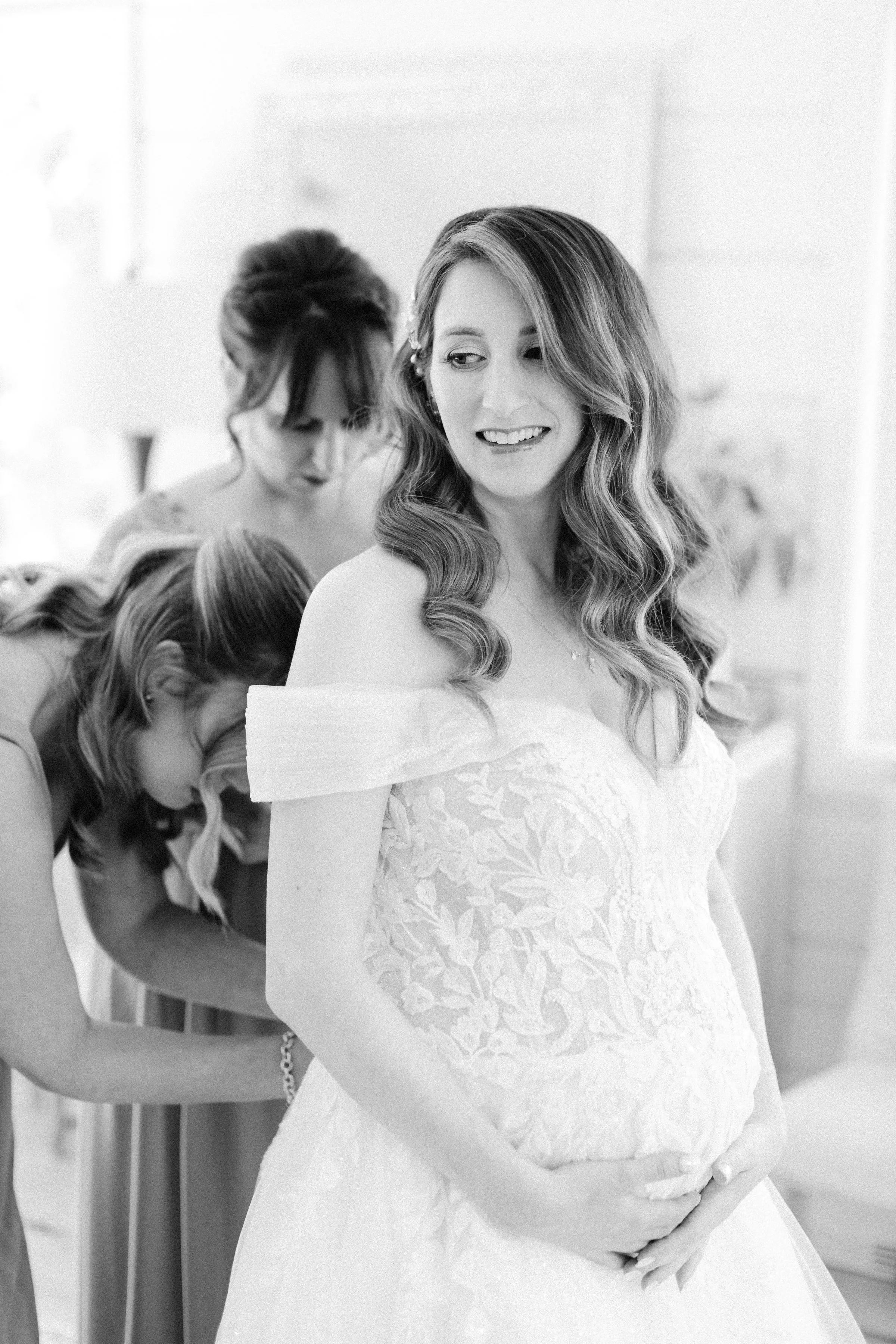 A bride with wavy hair wearing a lace wedding dress holds her belly while two women, likely her bridesmaids, help her with dresses in the background, all in a bright, indoor setting.