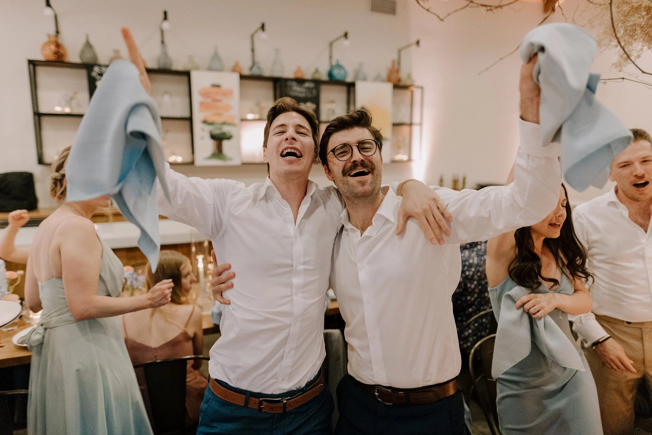 Two men in white shirts celebrating and smiling with arms around each other in a lively restaurant or party setting with other people in the background.
